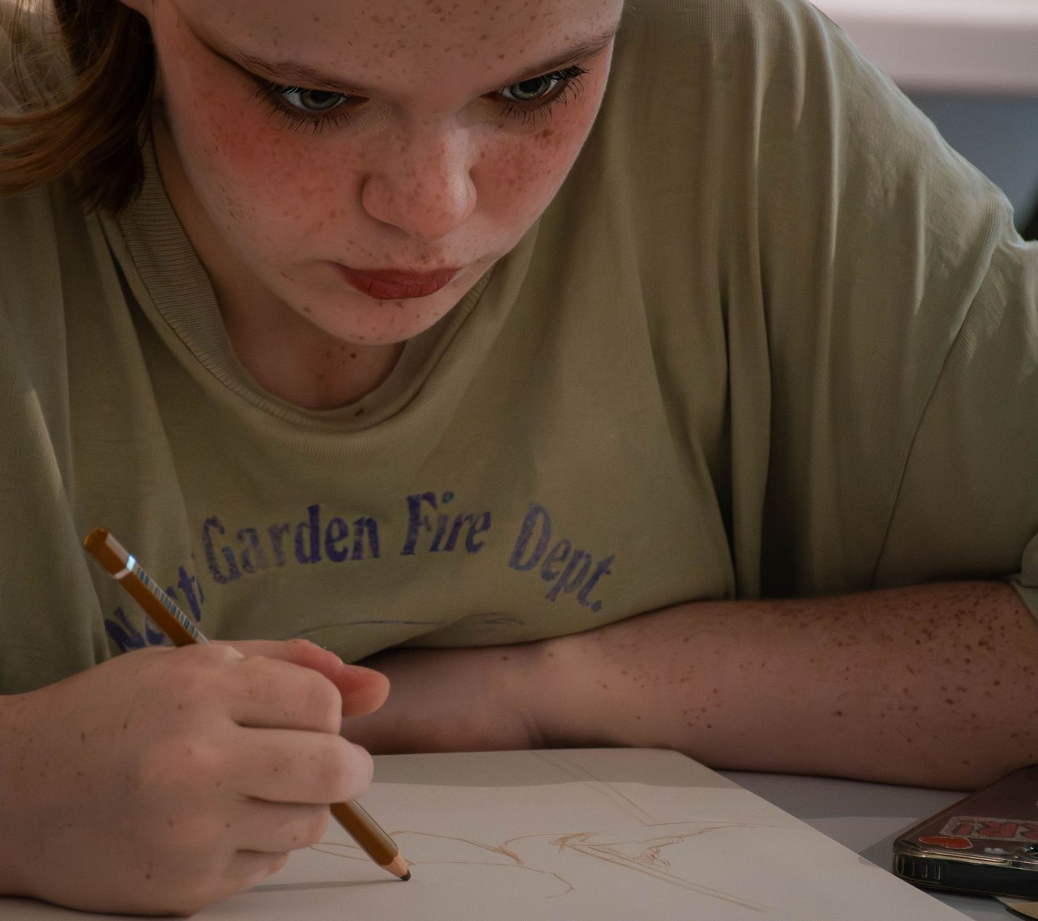 A freckled young girl in a Garden Fire Dept shirt draws carefully with a pencil on white paper.