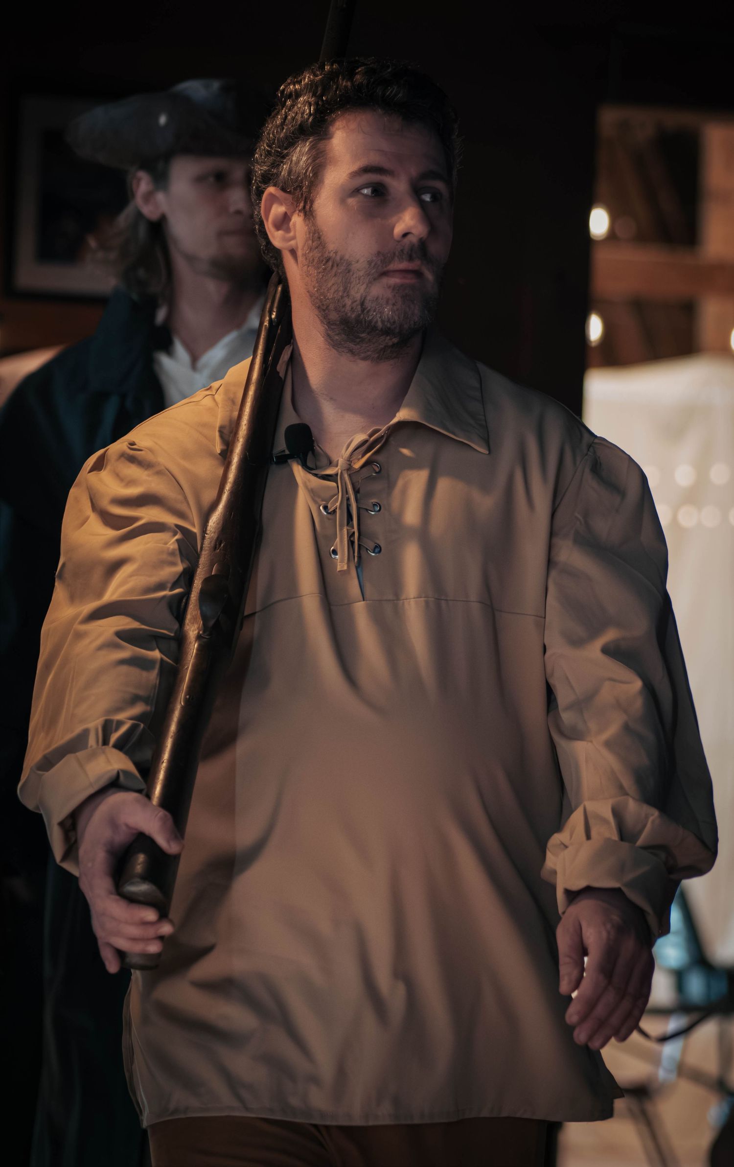 Man in colonial costume holding a musket over his shoulder in a dimly lit historical theater production.