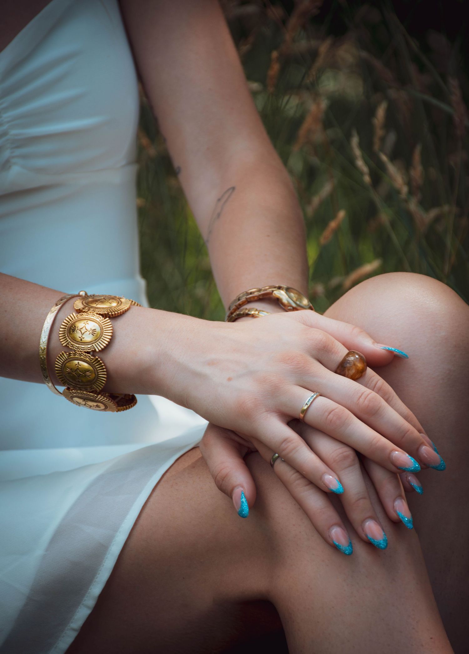 Woman wearing gold coin bracelets and turquoise-tipped nails with amber ring, hands resting on knee in white dress.
