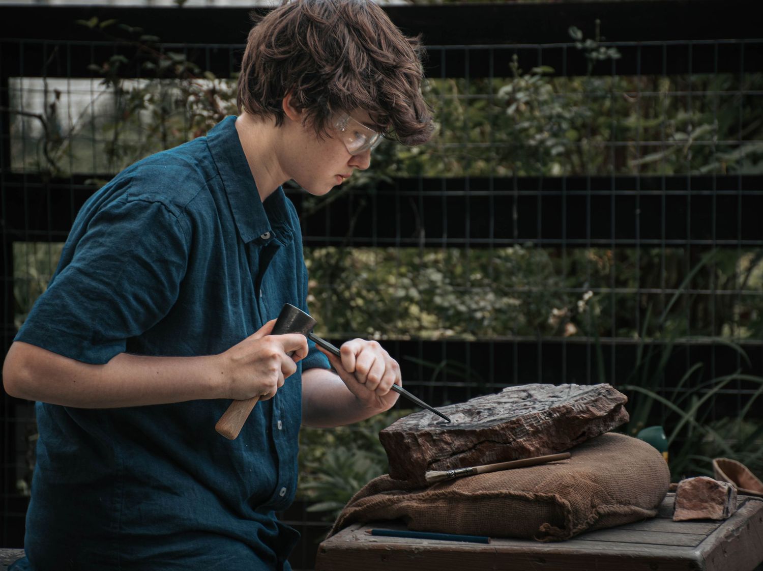 Young man wearing safety glasses carefully carving or sculpting wood outdoors with a chisel.