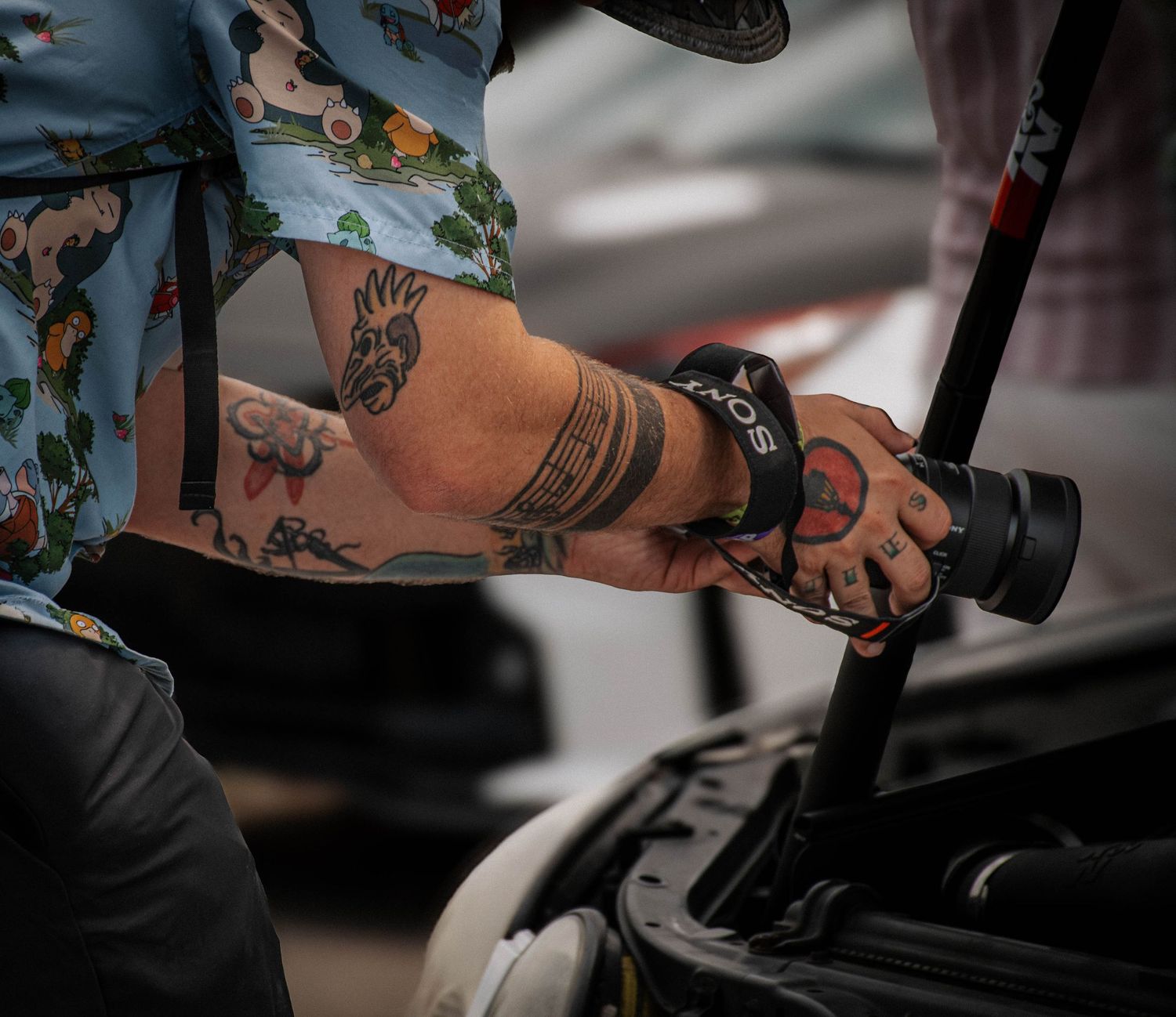 Tattooed arm holding a camera lens near a car engine, wearing a floral shirt and camera strap.