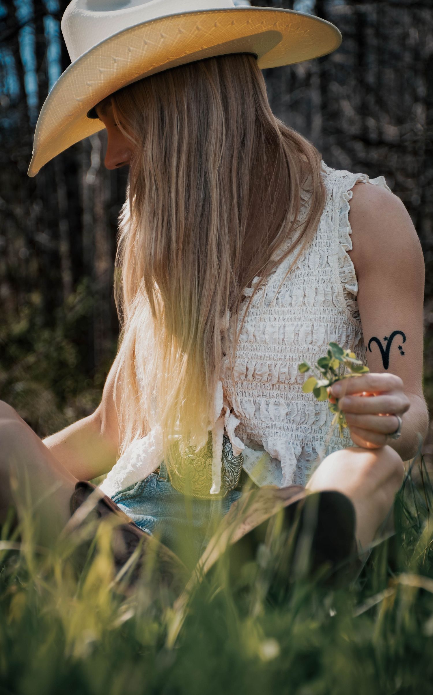 Blonde woman in cowboy hat and white lace top sits in grass holding wildflowers.