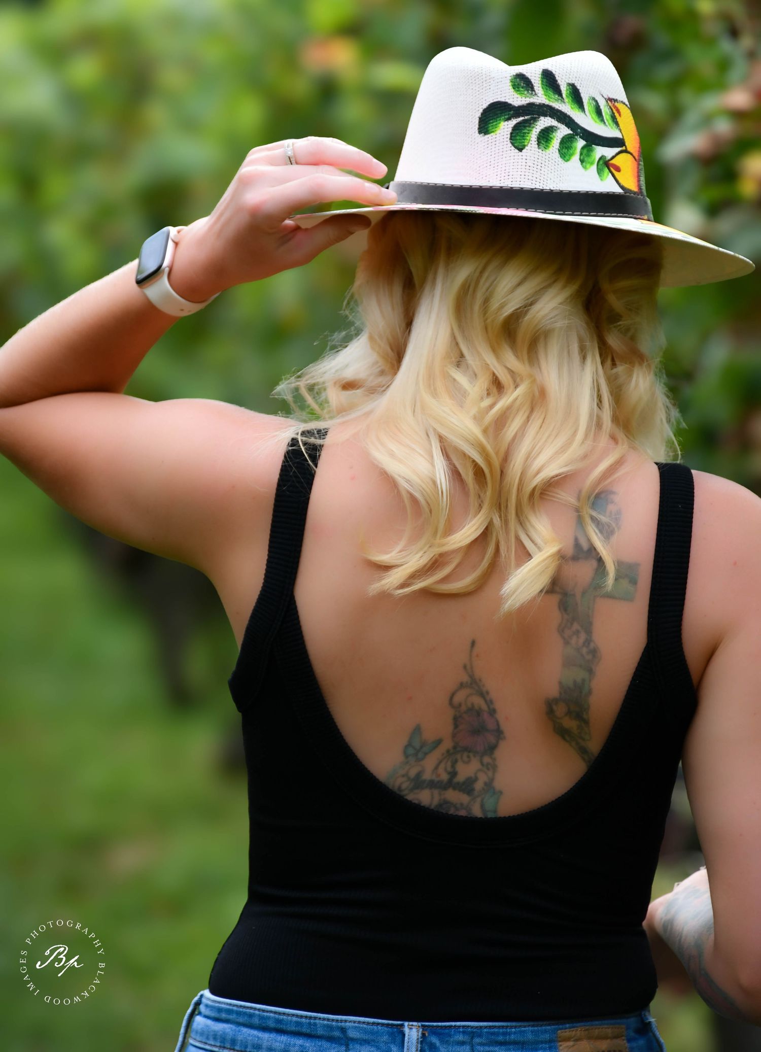 Blonde woman with back tattoos wearing painted green leaf fedora hat in lush vineyard setting.