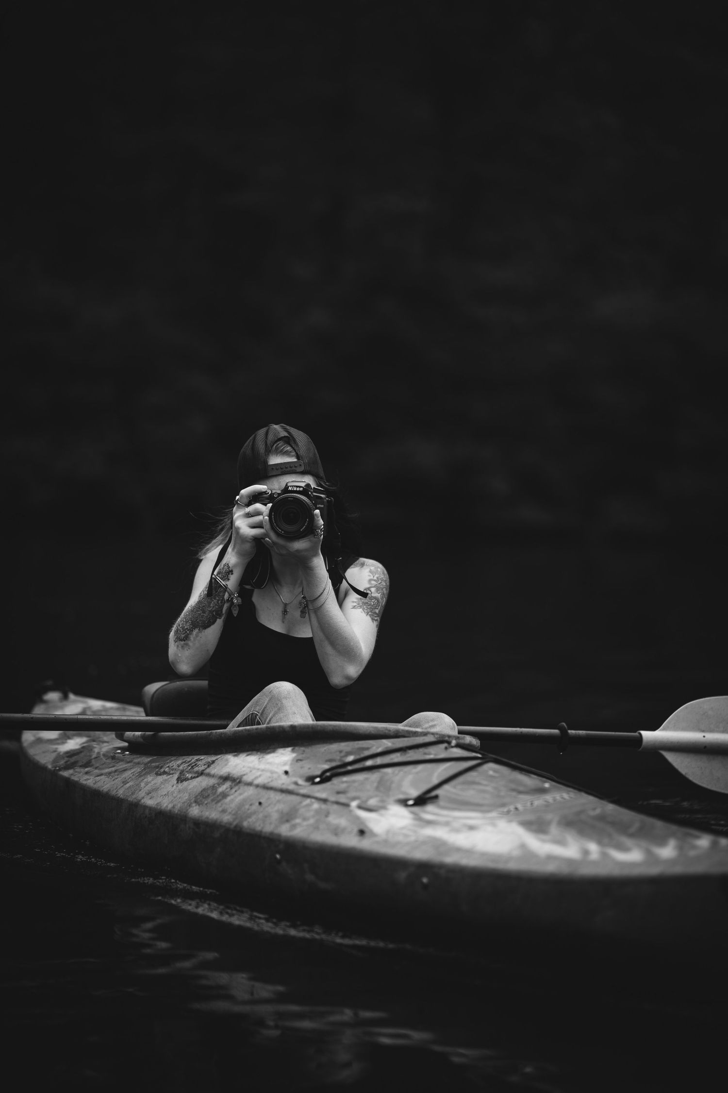 Black and white photo of a woman sitting in a kayak, holding a camera up to her face, with a dark water background.