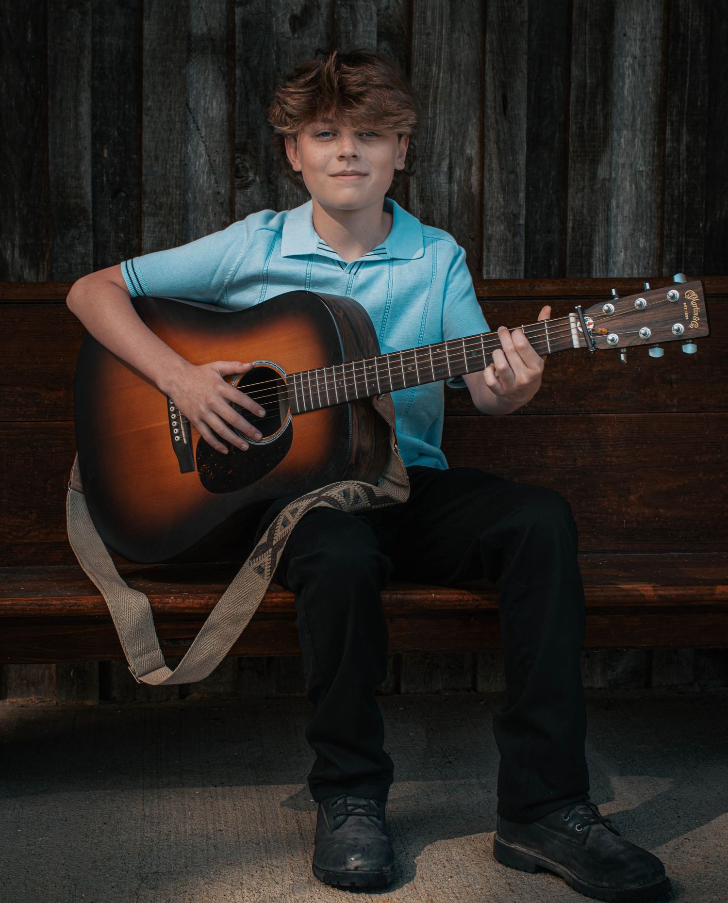 Young boy in blue polo shirt playing acoustic guitar while seated on a bench, smiling confidently.