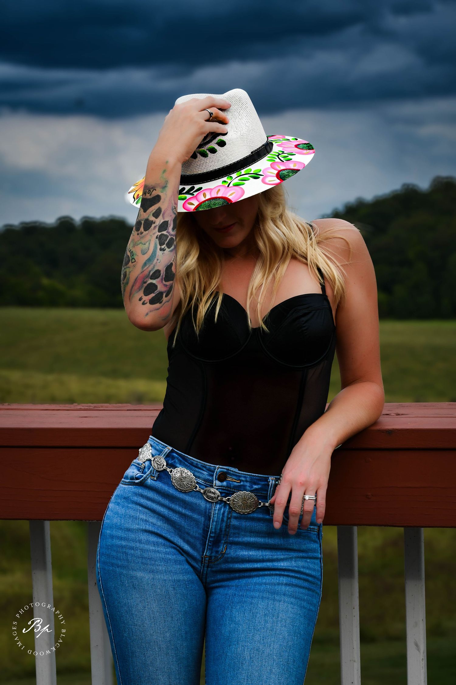 Blonde woman in black corset top and jeans holds floral hat on stormy outdoor background.