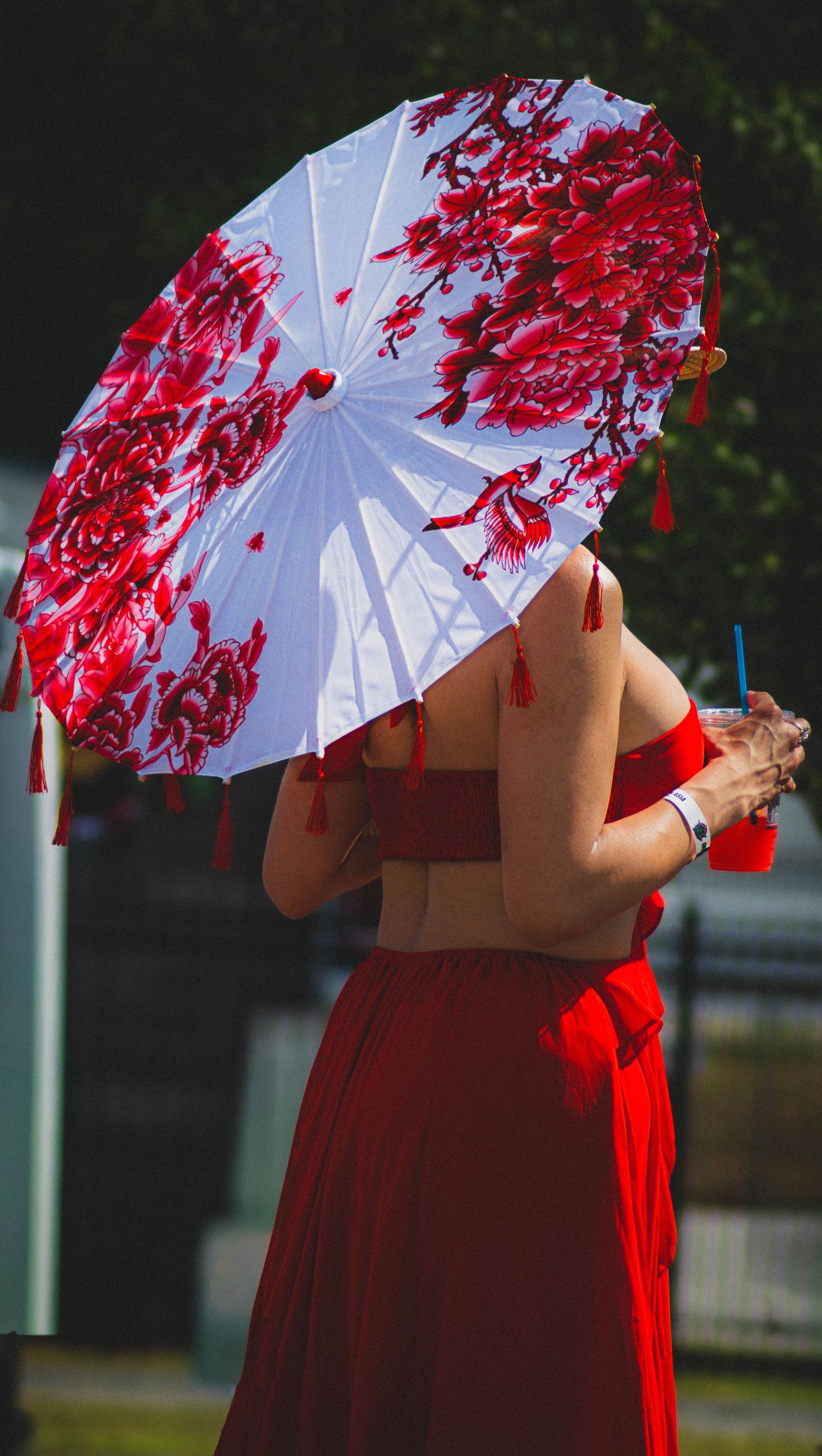 Woman in red dress holding a white parasol with red floral cherry blossom design and tassels outdoors.