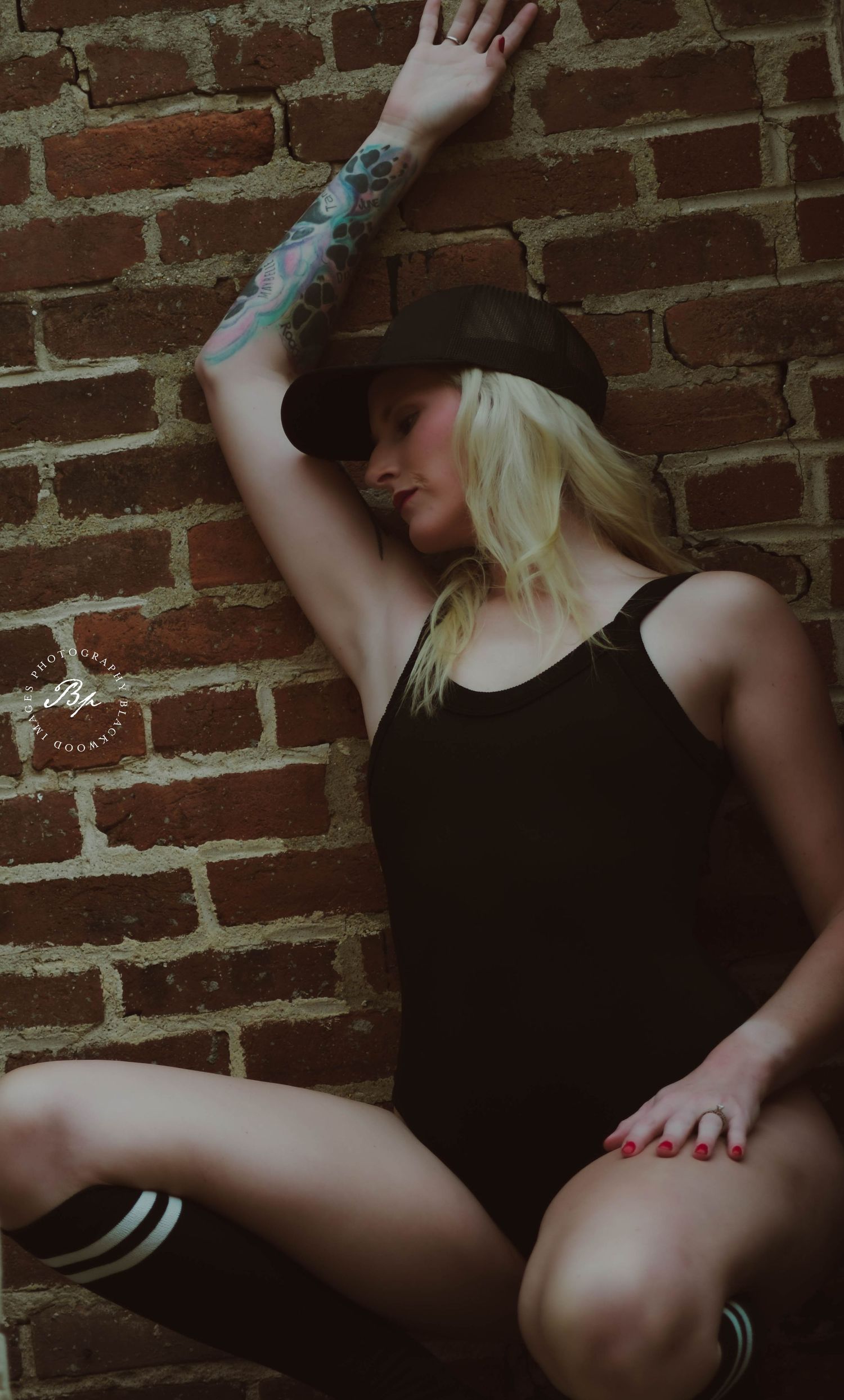 Blonde tattooed woman in black bodysuit and cowboy hat poses against rustic brick wall.