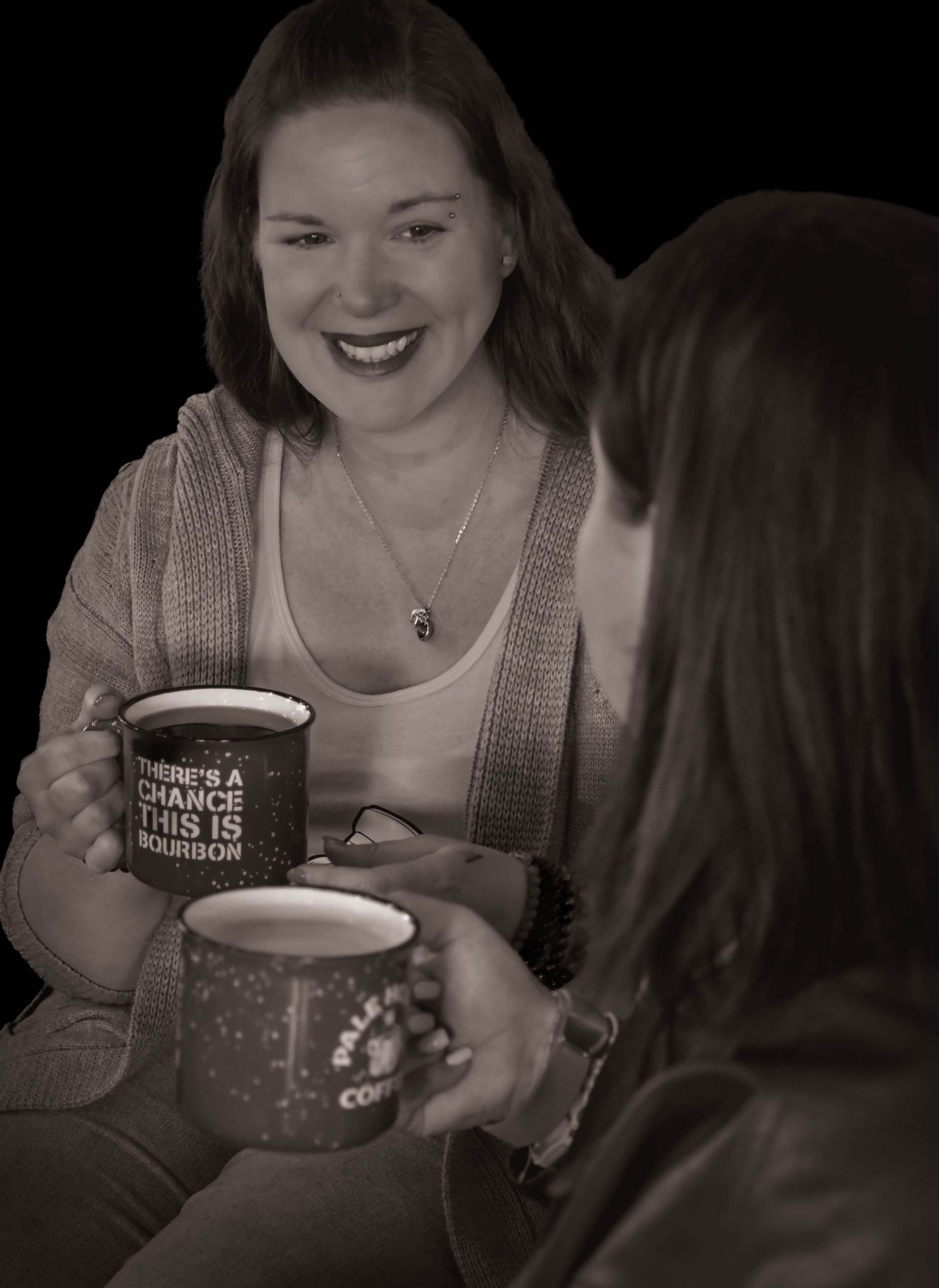 Two women laugh together holding There's a Chance This Is Bourbon mugs in sepia photo.