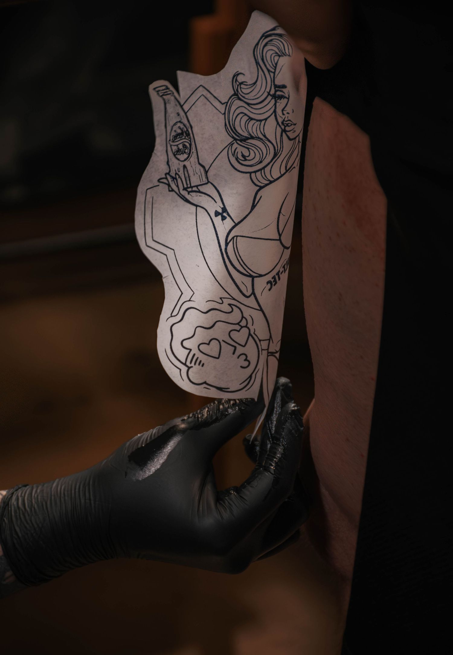Tattoo artist in black gloves holding a detailed stencil design against a client's arm in a tattoo studio.