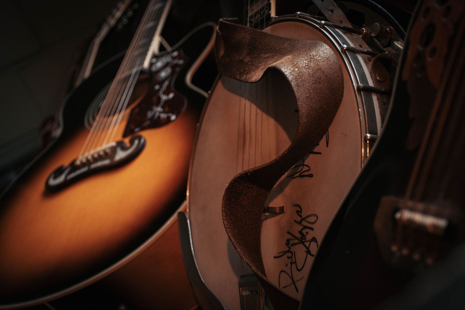 Close-up of vintage acoustic guitars and mandolin with signatures, showcasing warm sunburst finishes in moody lighting.