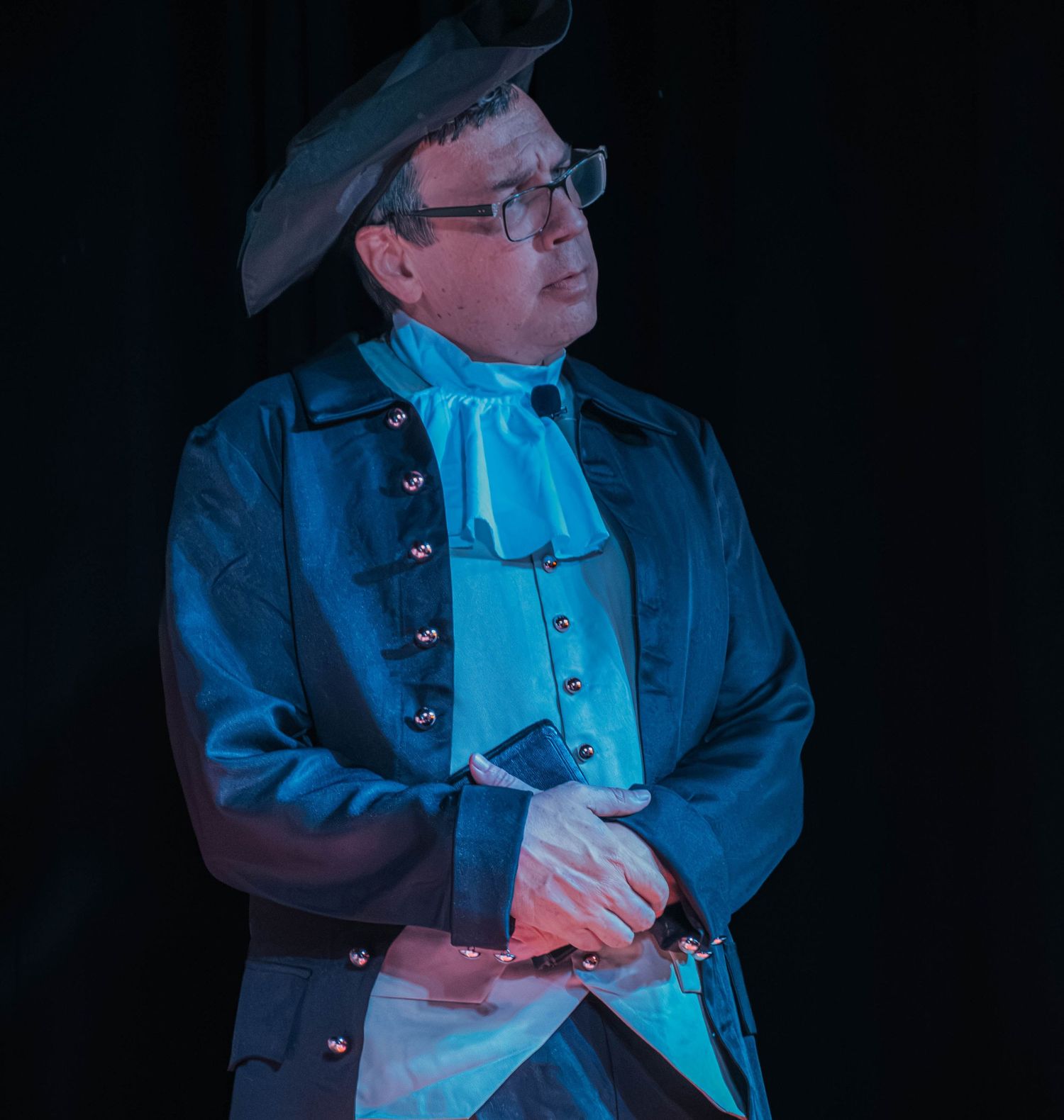Man in colonial-era costume with tricorn hat and blue coat poses dramatically on dark stage.