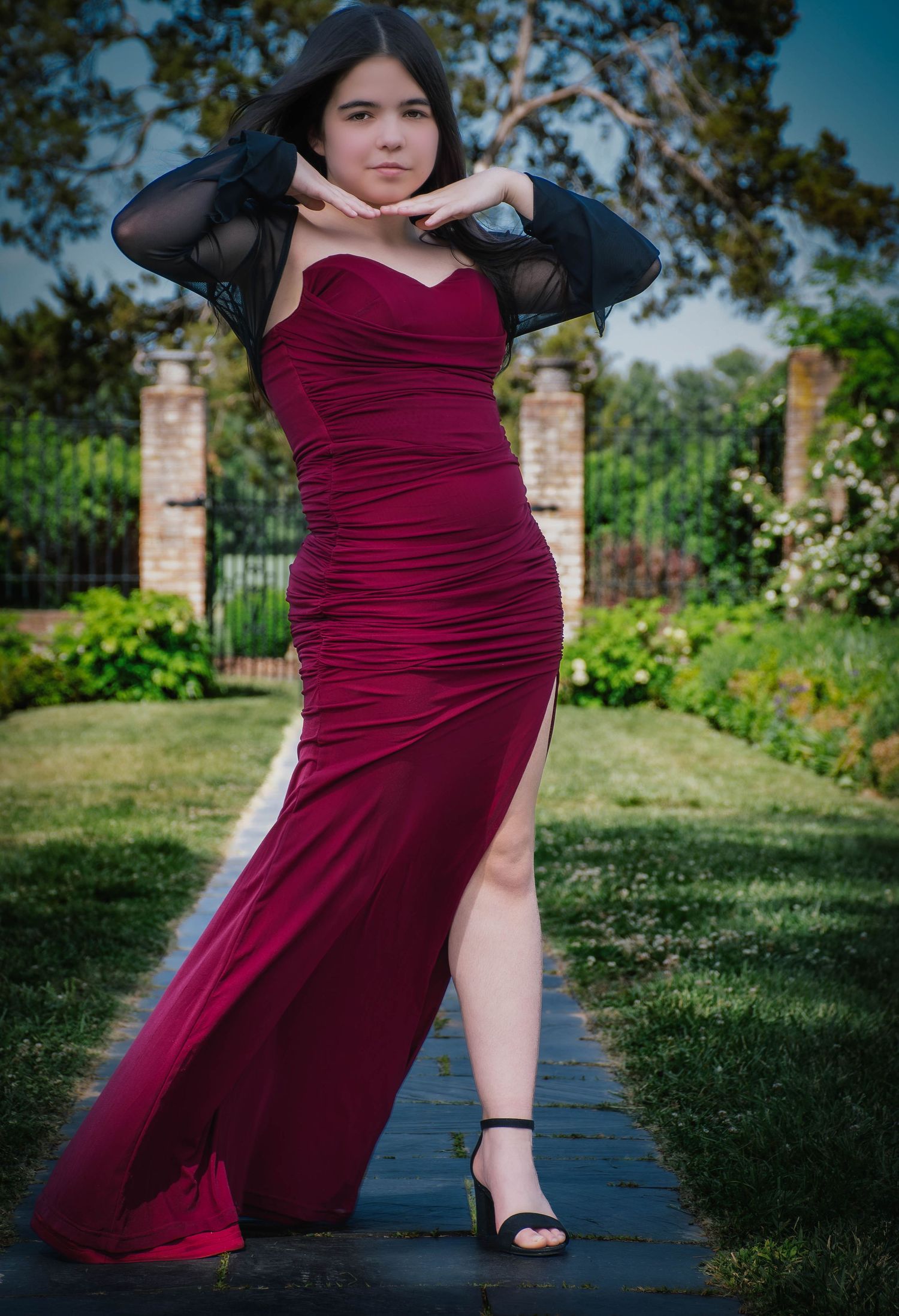 Young woman posing in red ruched formal gown with black sheer sleeves and high slit in garden setting.