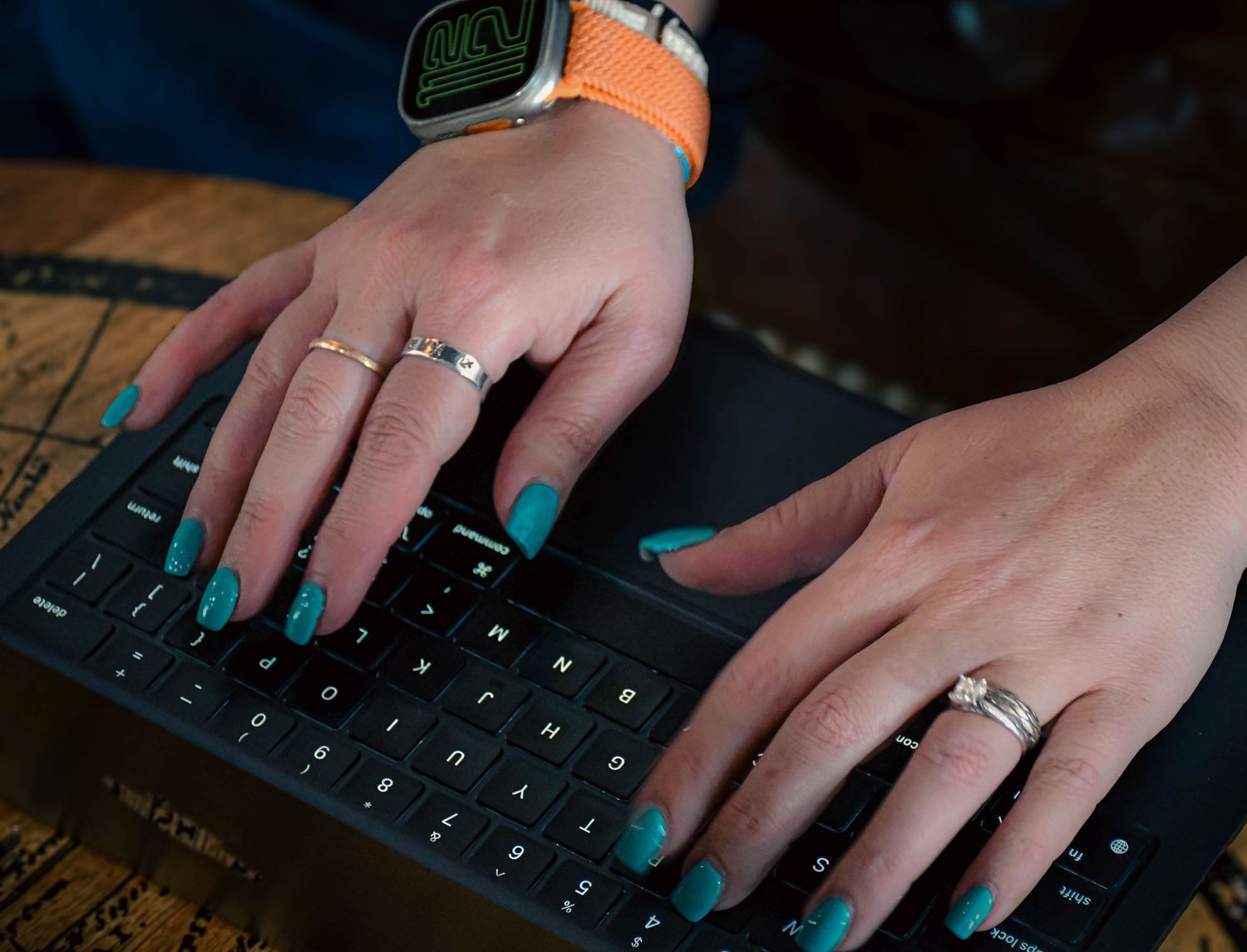Close-up of hands with teal nails and rings typing on a dark laptop keyboard with an Apple Watch.
