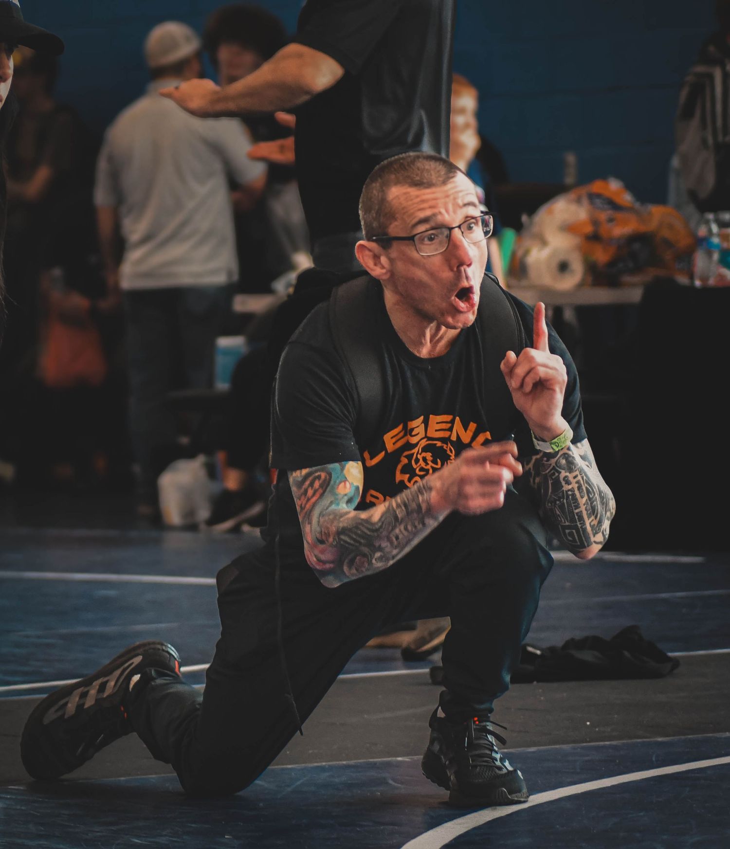 Tattooed wrestling coach kneeling on mat, pointing finger and coaching during a wrestling tournament.