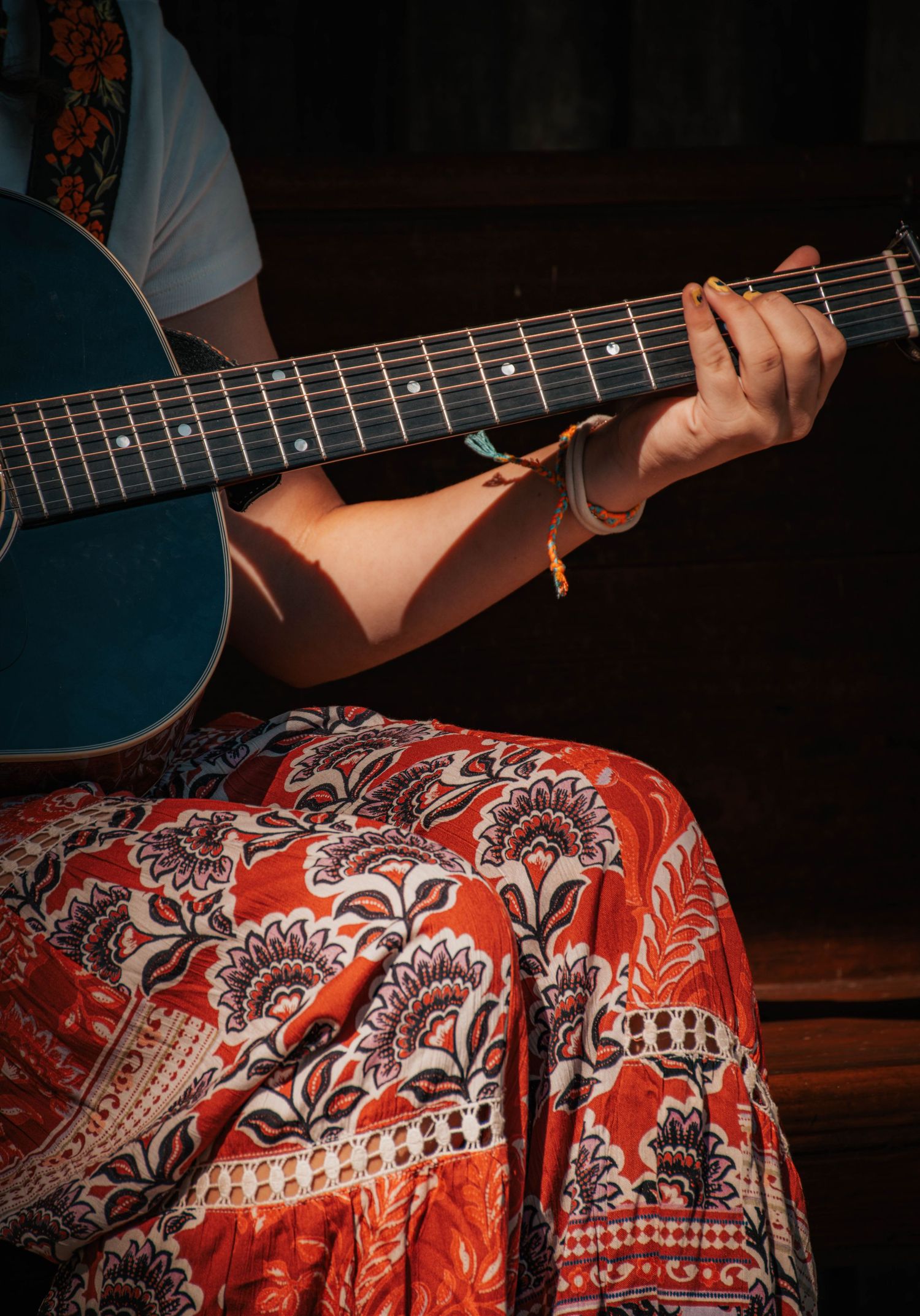 Person playing a blue acoustic guitar wearing a floral red skirt and bracelet, bathed in warm sunlight.