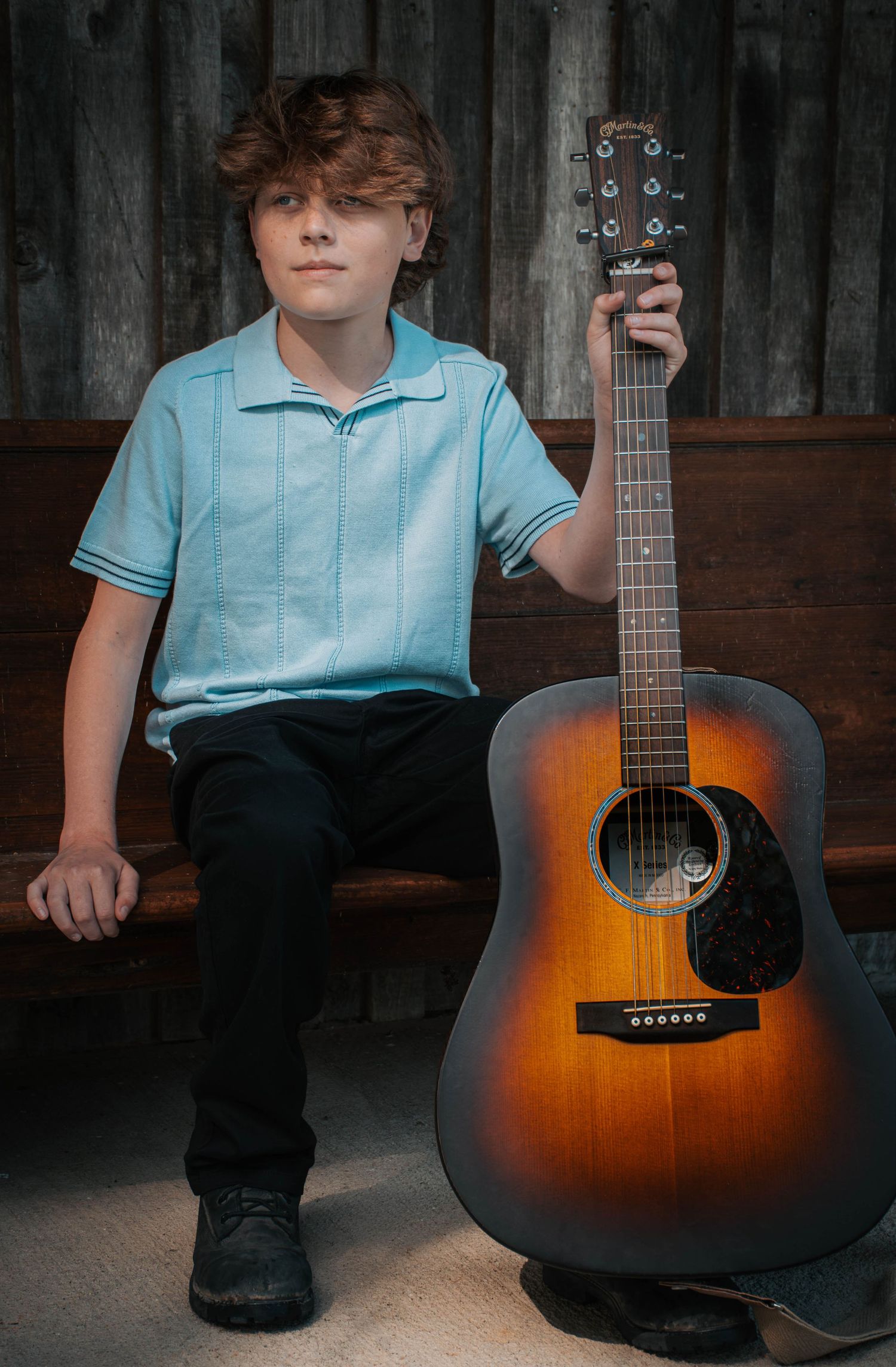 Young boy in light blue shirt holding acoustic guitar while seated on bench against rustic wooden wall.