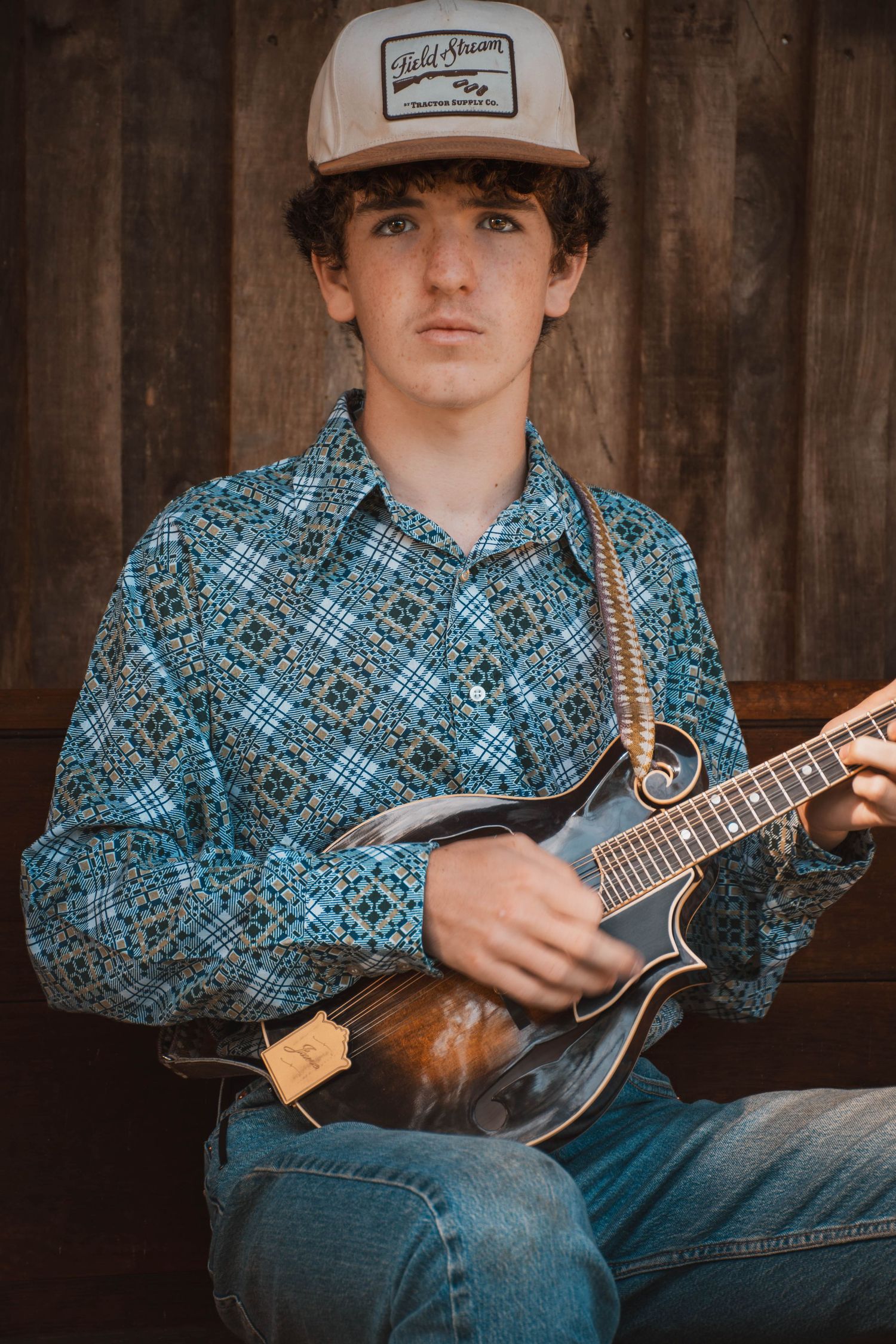 Young man in plaid shirt and trucker hat playing mandolin while seated against rustic wood wall.