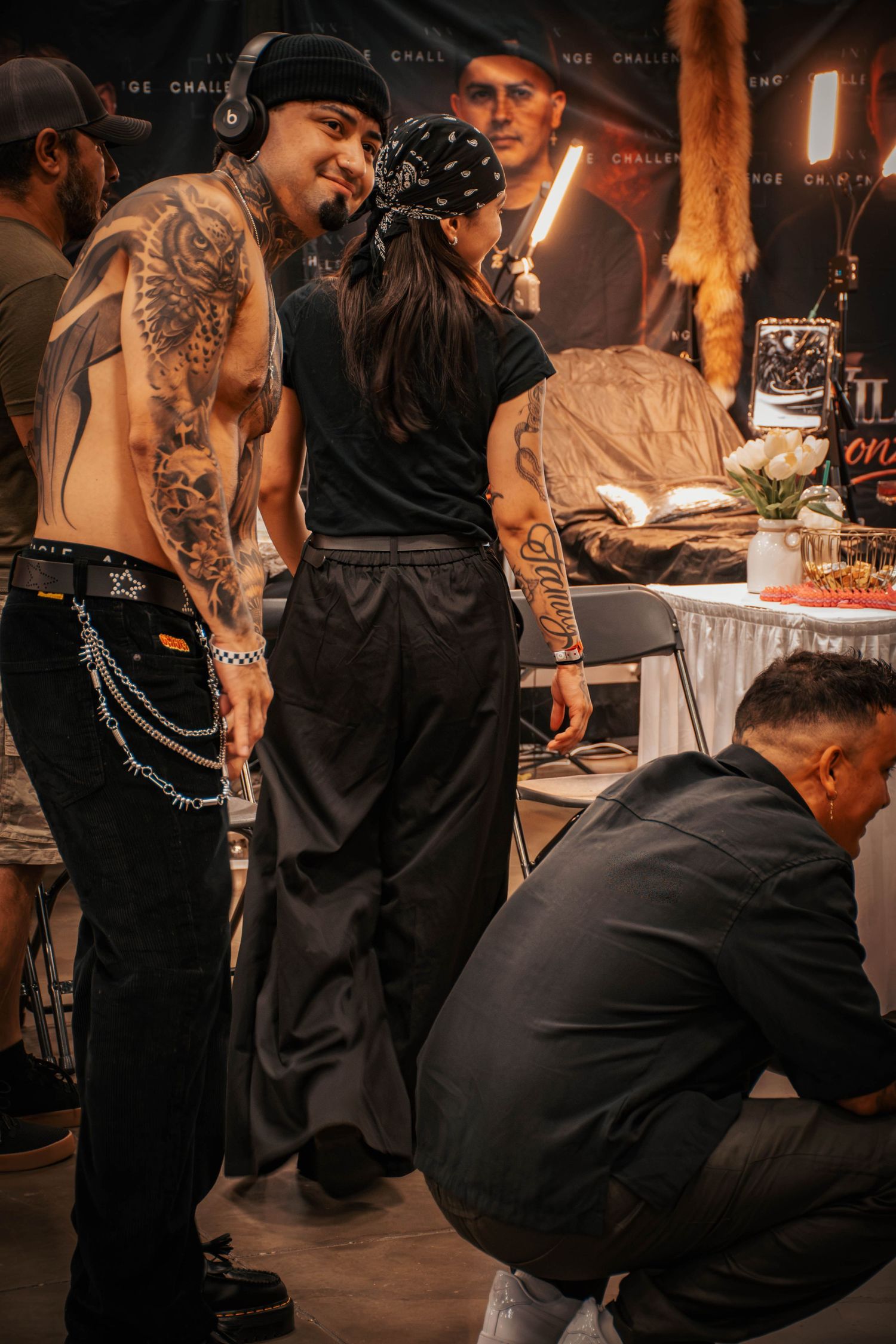 Tattooed man and woman interact at a tattoo convention event with other attendees in the background.