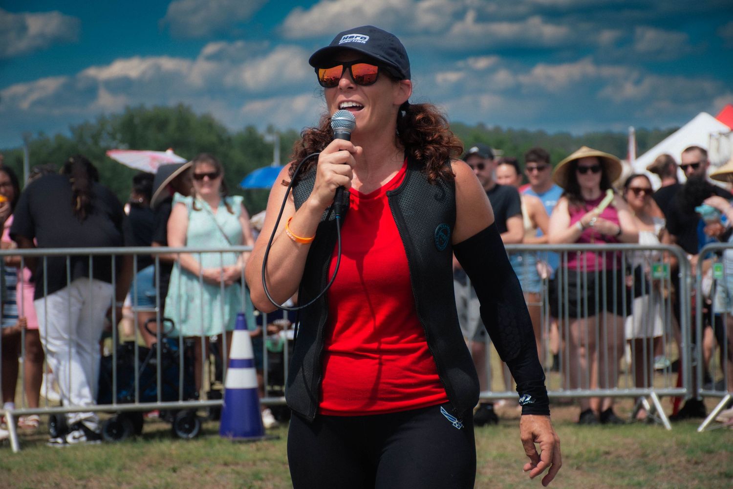 Woman in red top and cap speaks into microphone at outdoor event with crowd behind metal barriers.