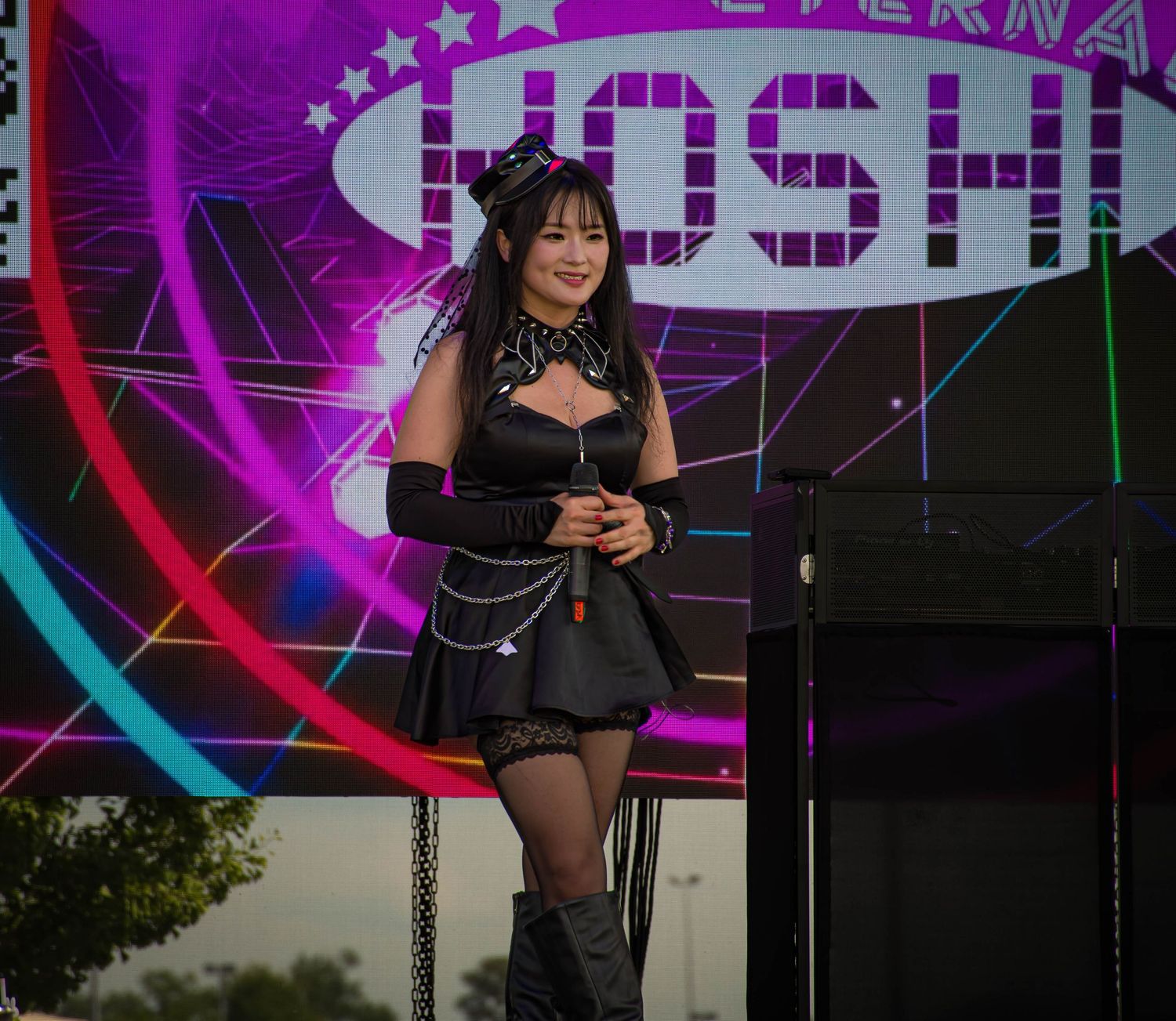 Woman in black gothic outfit performing on stage with colorful neon lights and Hoshi sign backdrop.
