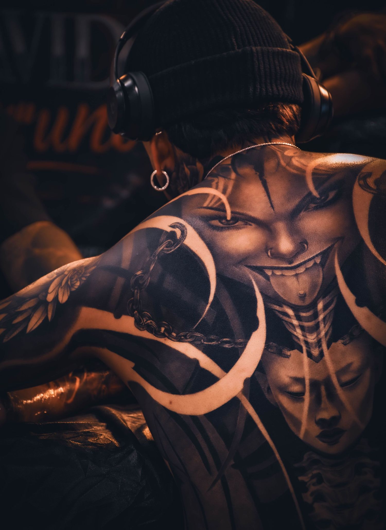 Person with elaborate black and grey back tattoo featuring a sinister face design, wearing headphones and beanie.