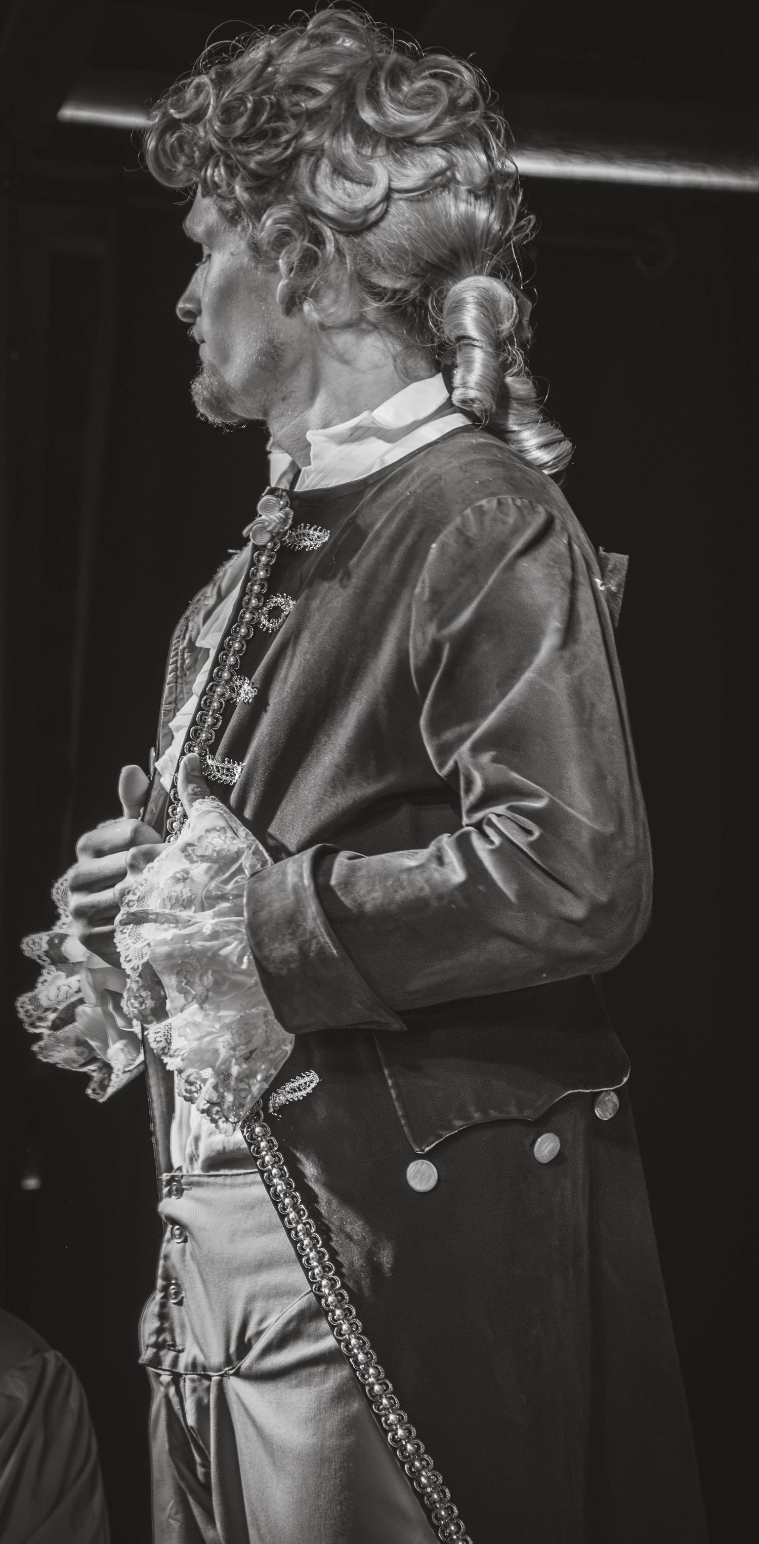Black and white photo of a man in 18th-century costume with ruffled shirt and ornate jacket, posing in profile.