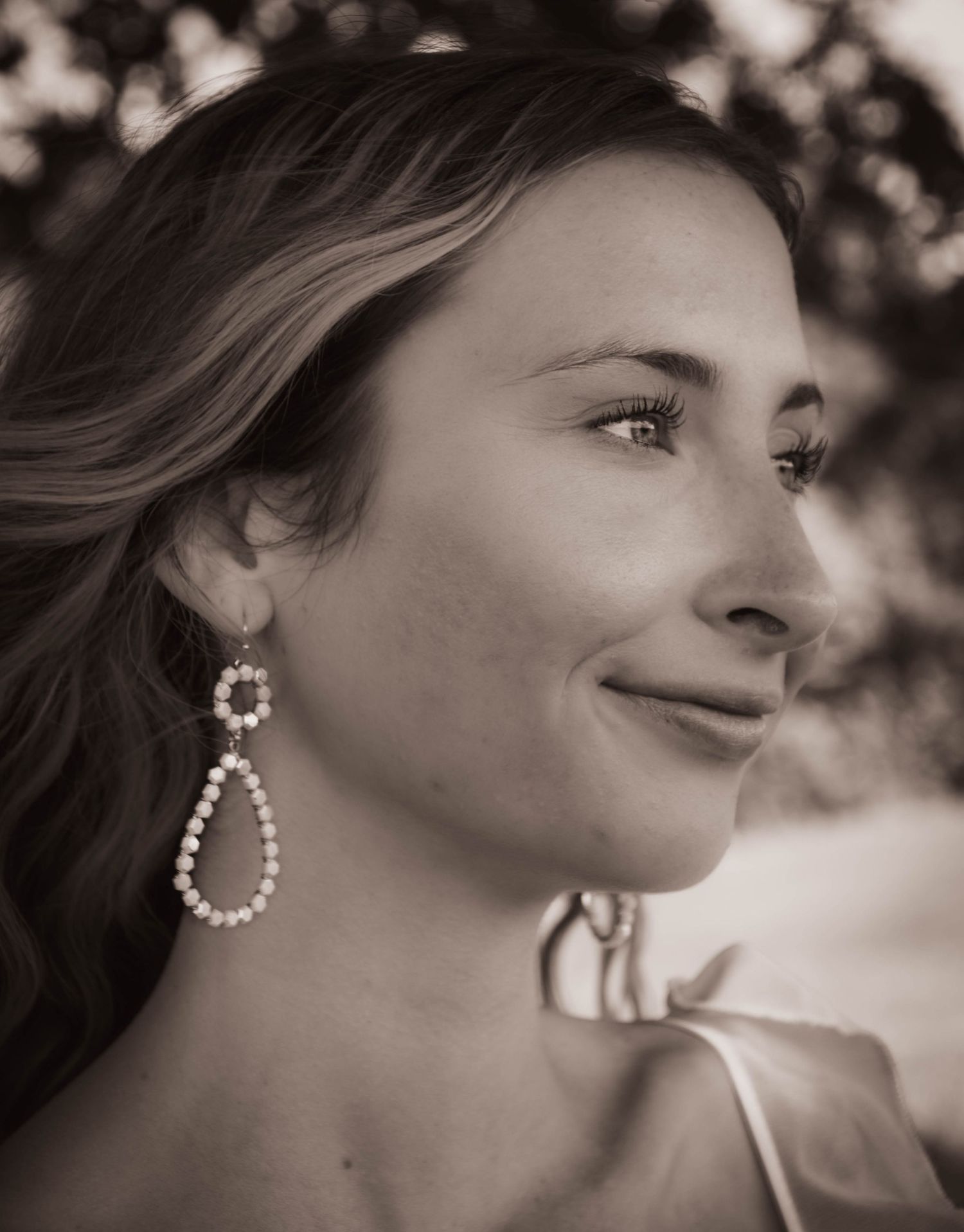 Sepia portrait of a smiling young woman with long hair wearing elegant pearl drop earrings outdoors.