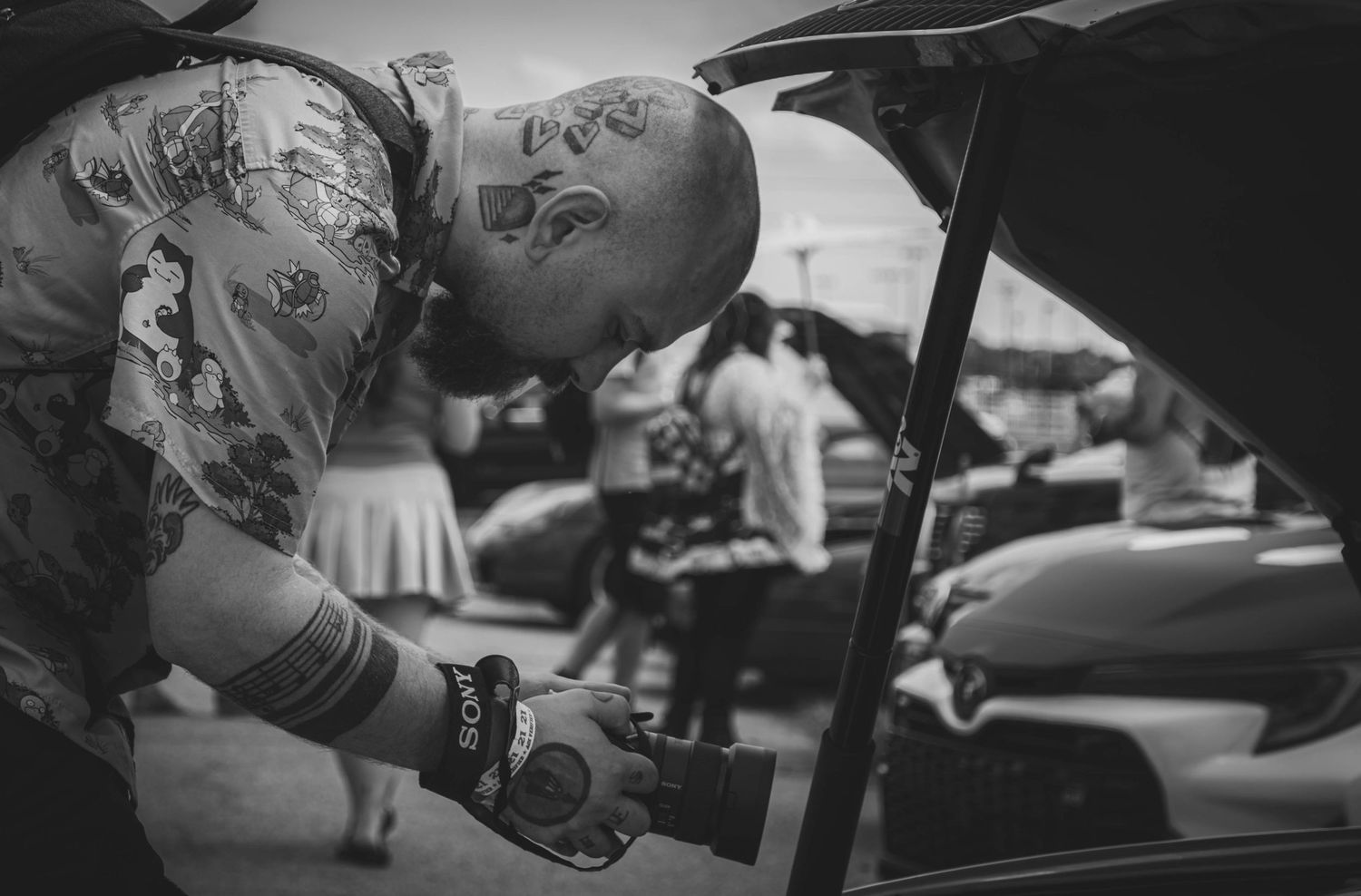 Bald tattooed man leaning over a car engine at an outdoor auto show in black and white photo.