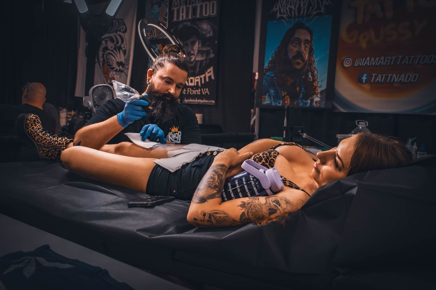 A tattoo artist applies ink to a client lying on a table in a professional tattoo studio with artwork on the walls.