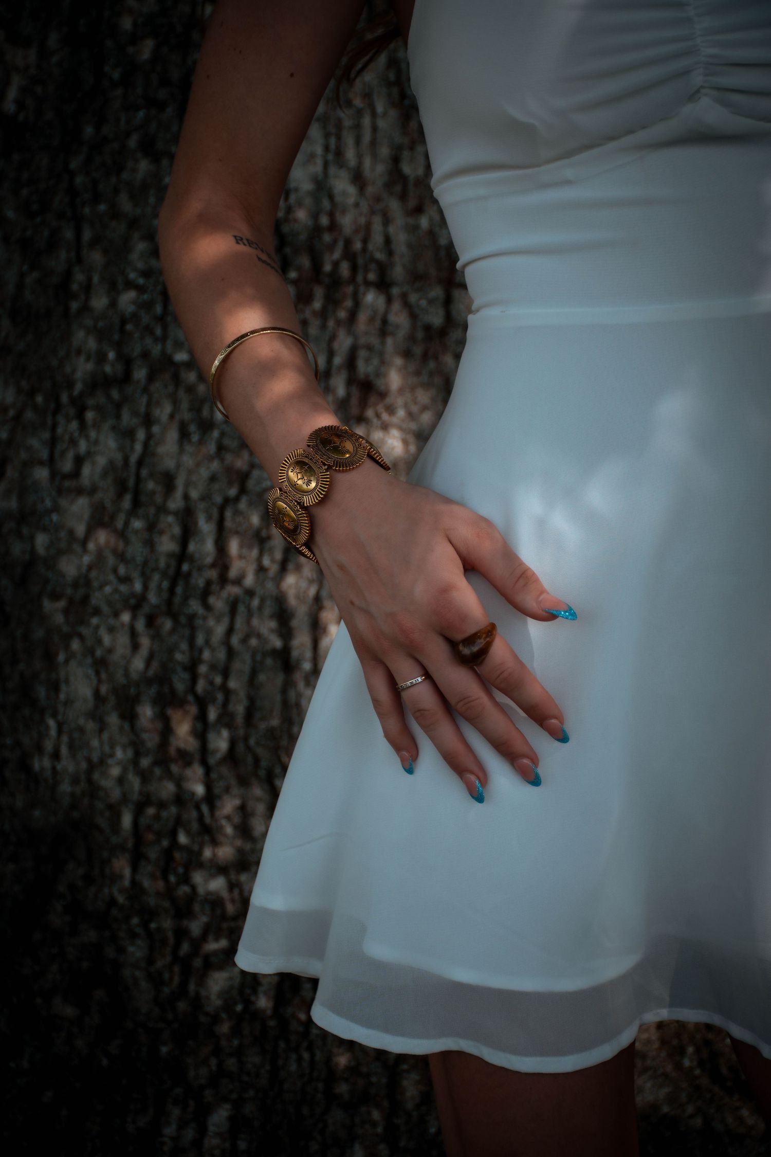 Woman in white dress wearing gold bracelet and ring with blue nail polish, posing near tree bark.