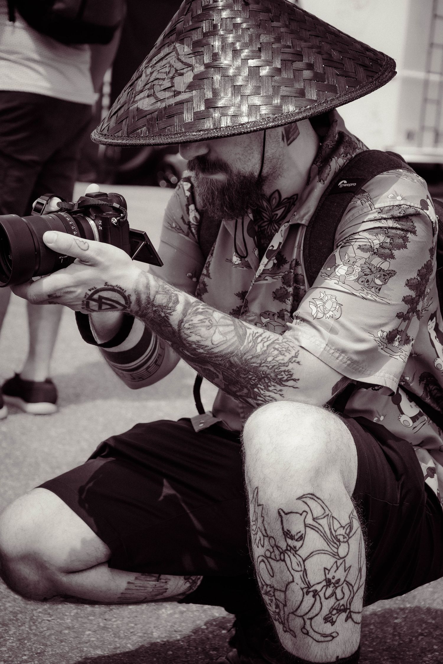 Tattooed bearded man wearing Asian conical hat sits cross-legged outdoors reviewing photos on DSLR camera in black and white.