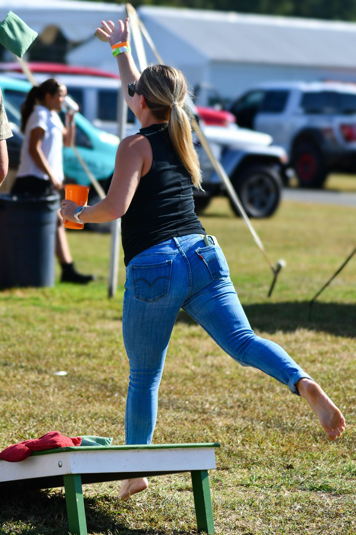 Blonde woman in black top and jeans tosses bean bag while playing cornhole at outdoor event.