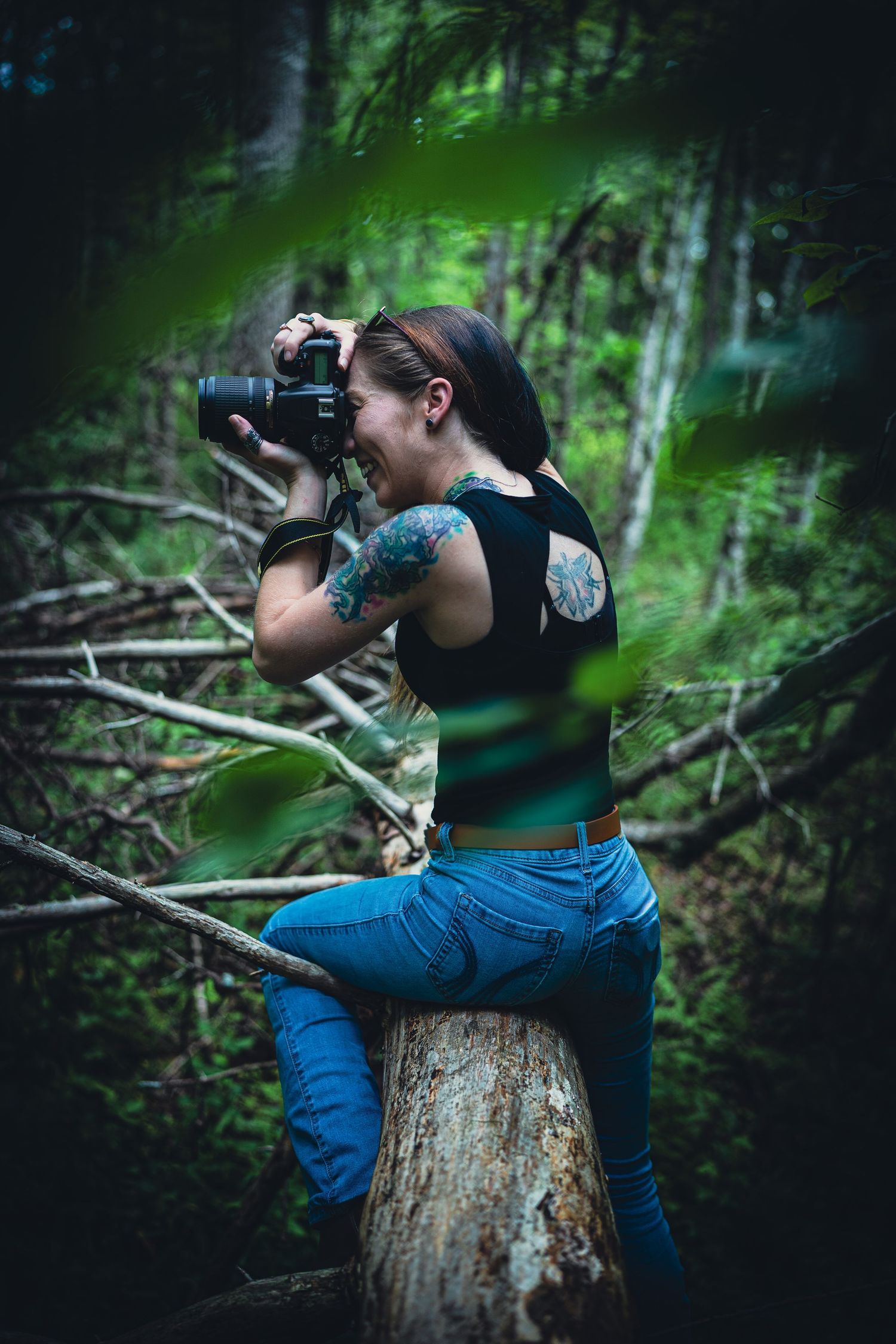 Tattooed woman photographer sitting on tree stump in forest, capturing photos with DSLR camera amid lush green woodland.