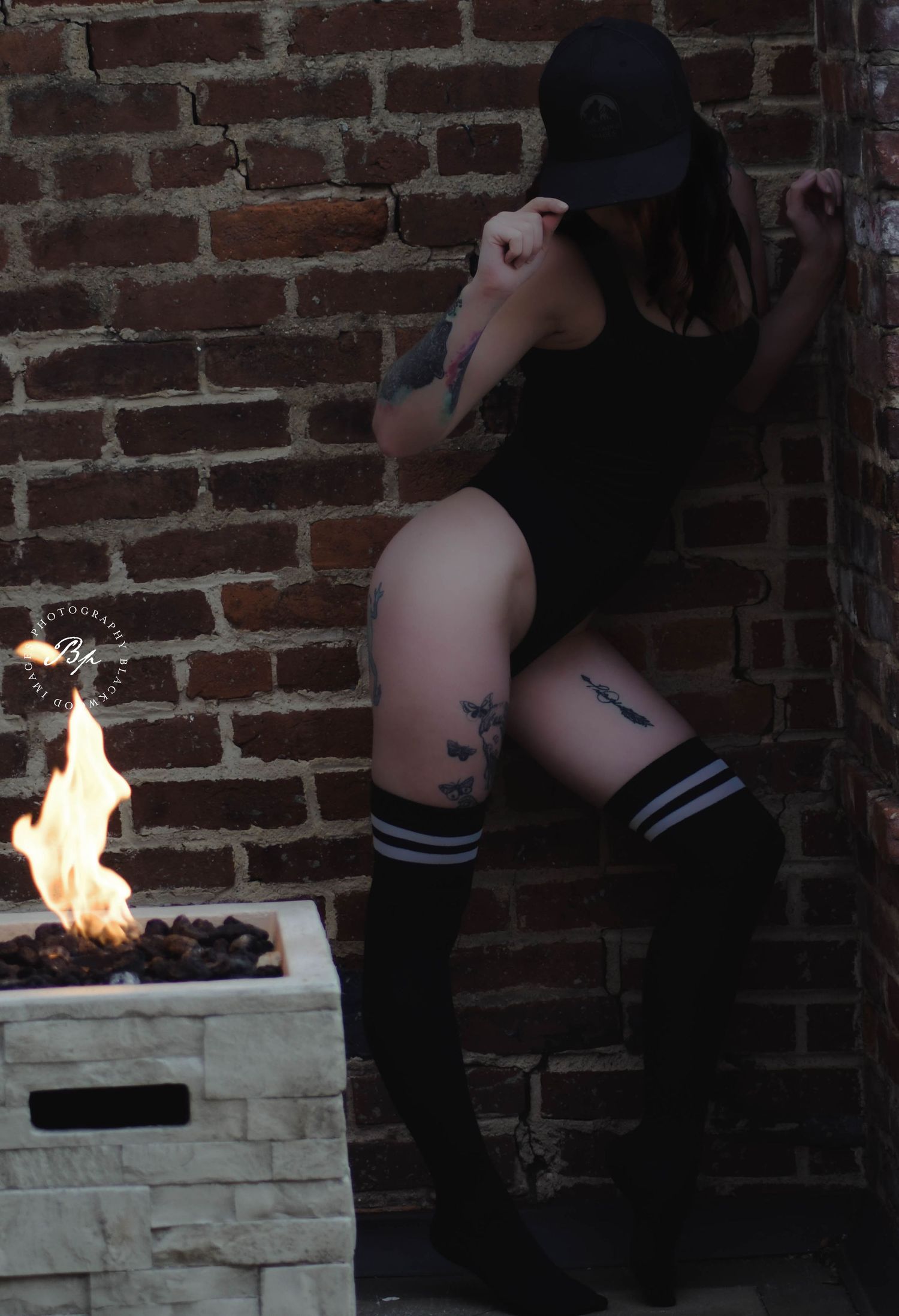 Tattooed woman in black bodysuit and knee-high socks poses by brick wall near outdoor fire pit.