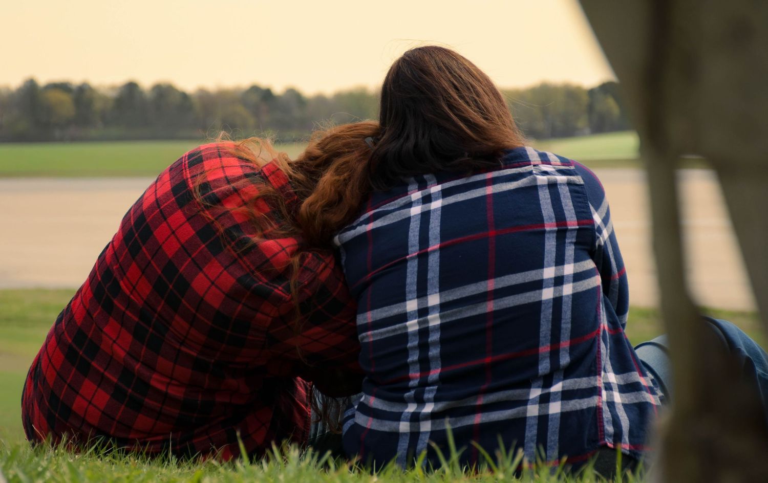 Two women in plaid shirts sitting together on grass, leaning on each other in a park setting.