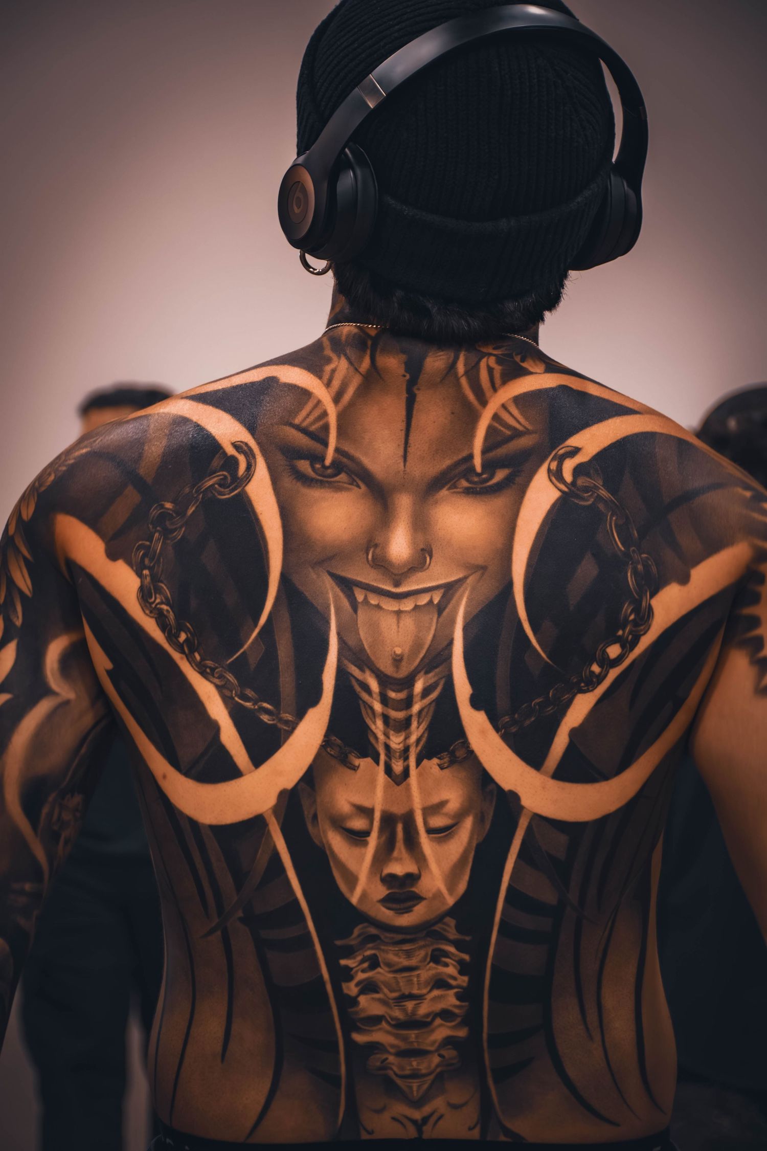 Man with intricate demon full-back tattoo wearing black beanie and headphones, showcasing detailed black ink body art.