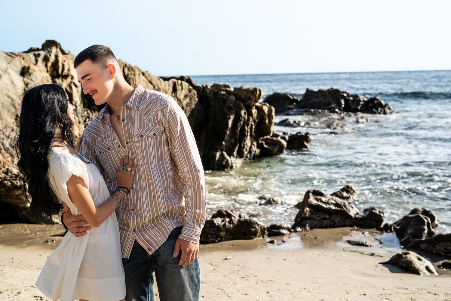 Romantic couple sharing a tender moment on a rocky beach with ocean waves nearby.