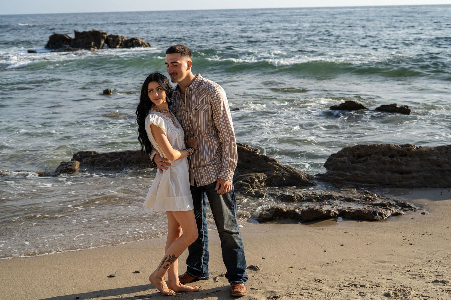 Couple posing together on a sandy beach with ocean waves and large rocks in background.