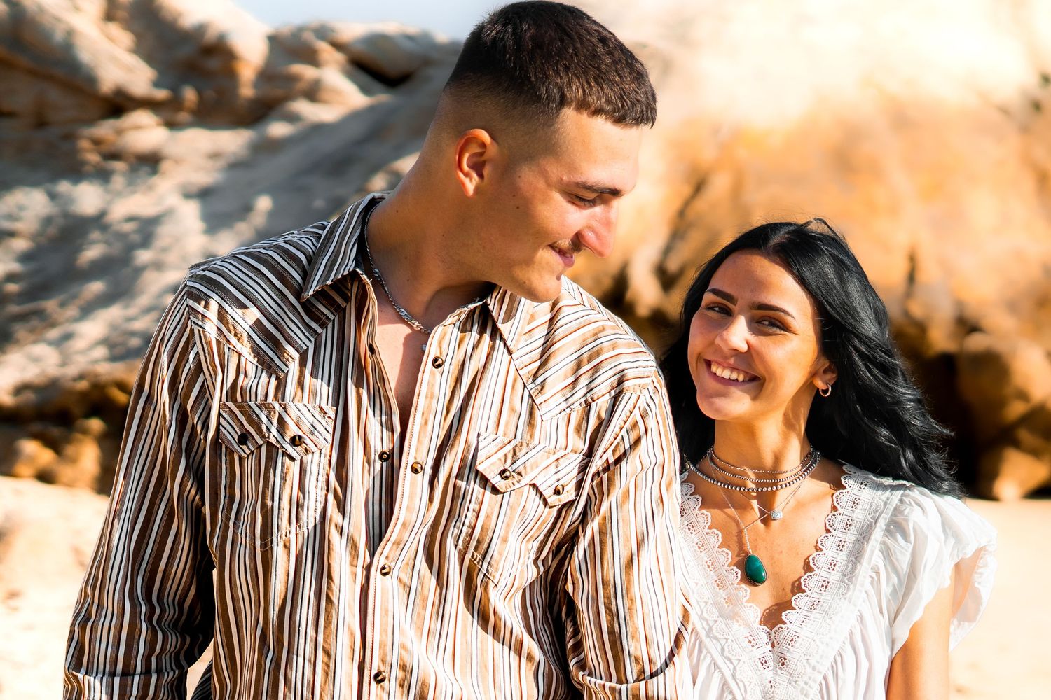 A smiling couple in casual attire poses outdoors near rocky terrain in warm golden light.