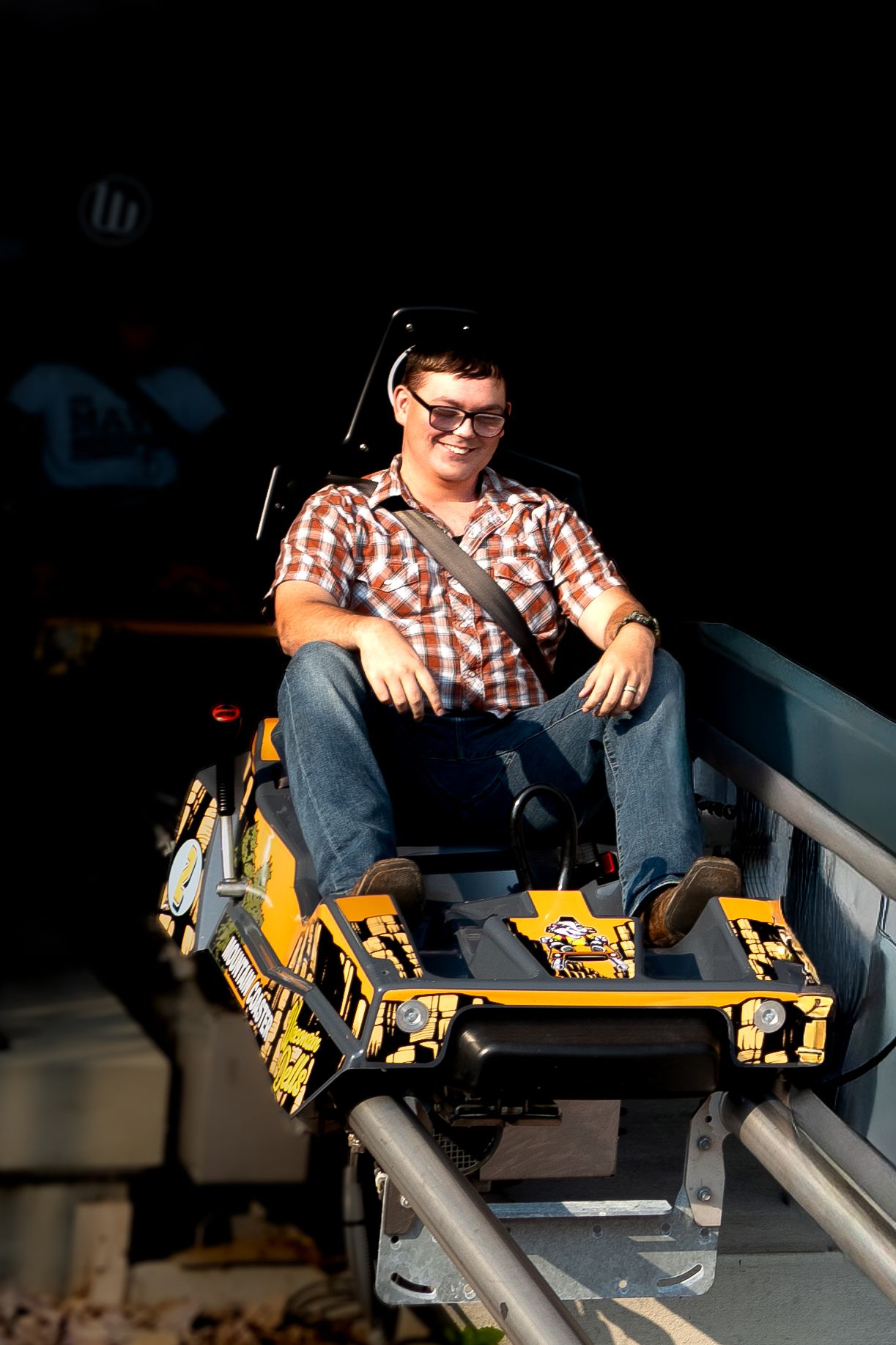 Young man wearing glasses and plaid shirt rides a yellow roller coaster car on a track at an amusement park.