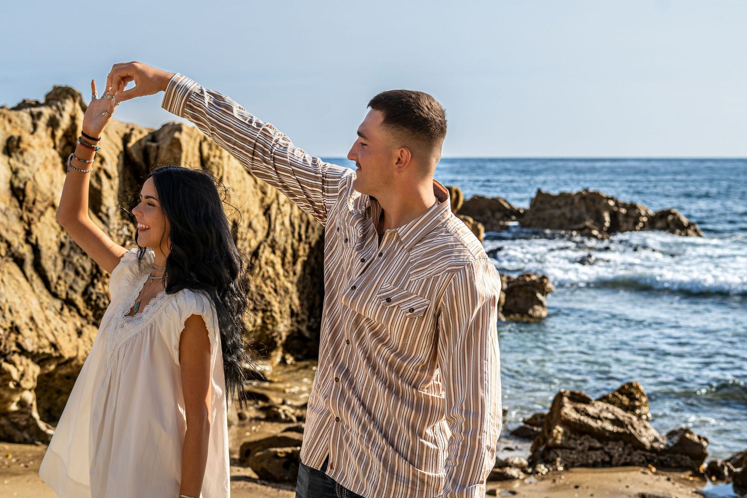 A couple dances romantically on a rocky beach, man spinning woman in white dress with ocean waves behind them.