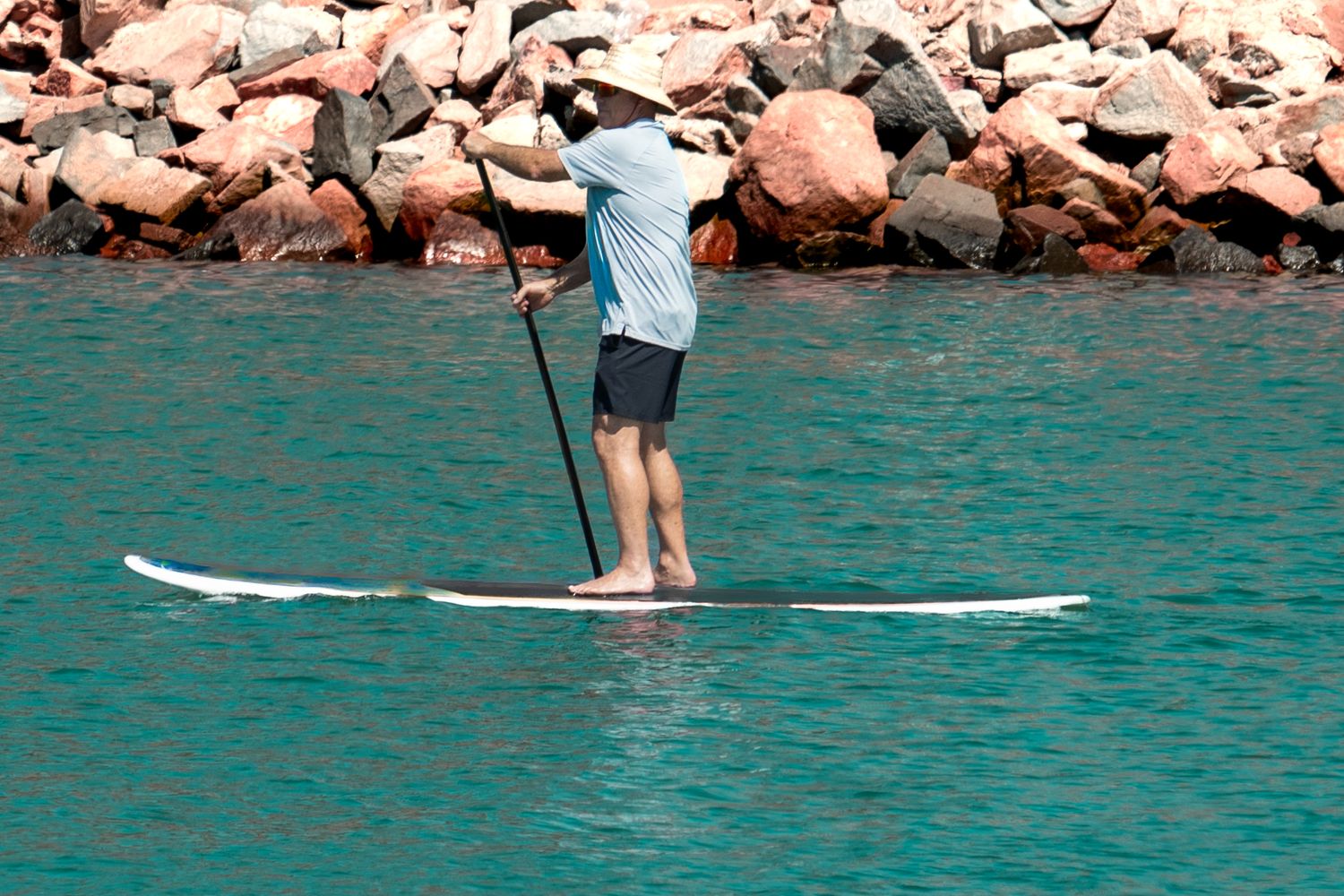 Man stand-up paddleboarding on calm turquoise water near a rocky shoreline.