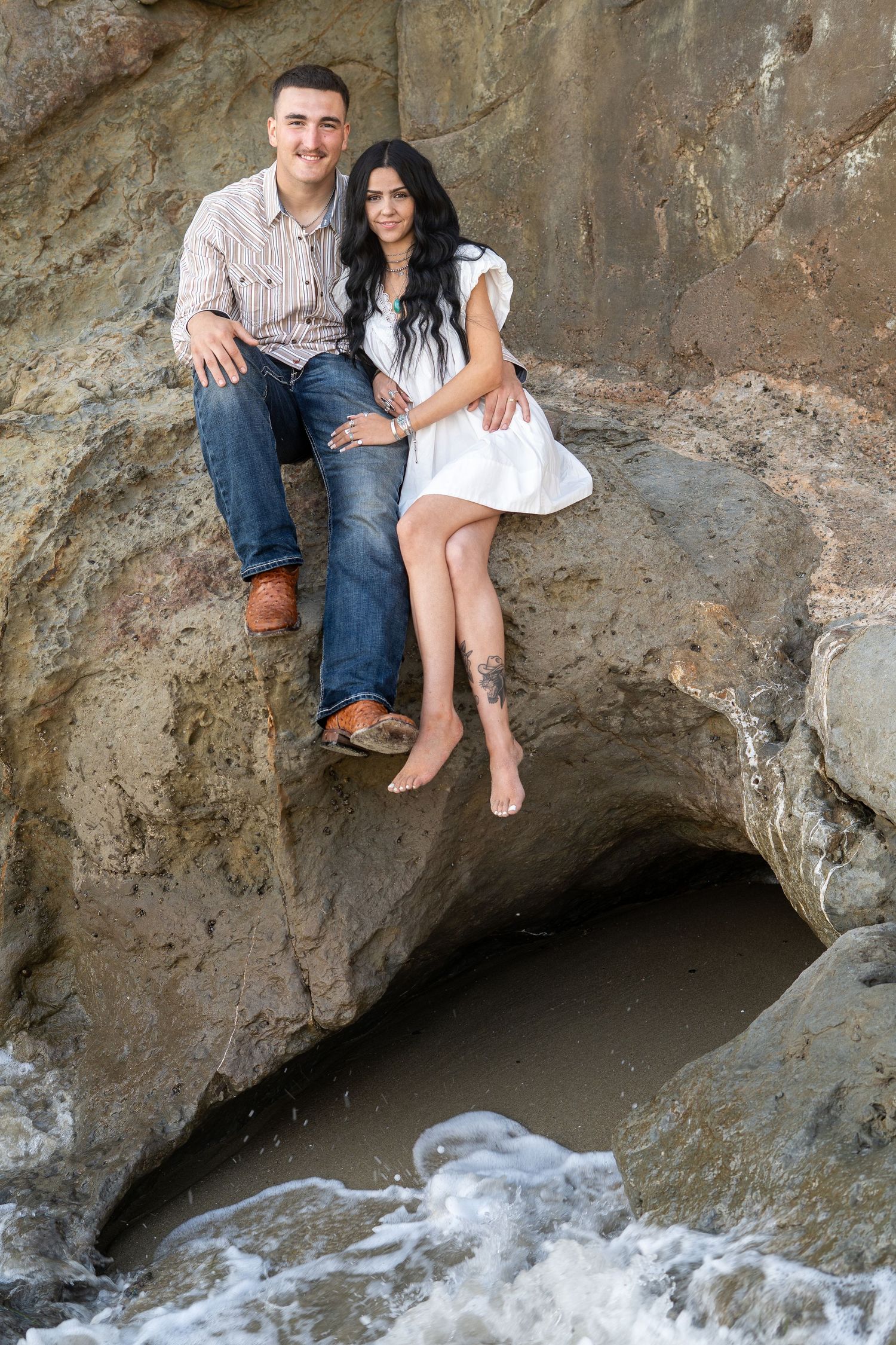 Couple sitting together on coastal rocks above ocean waves, woman in white dress with tattoo, man in jeans smiling.