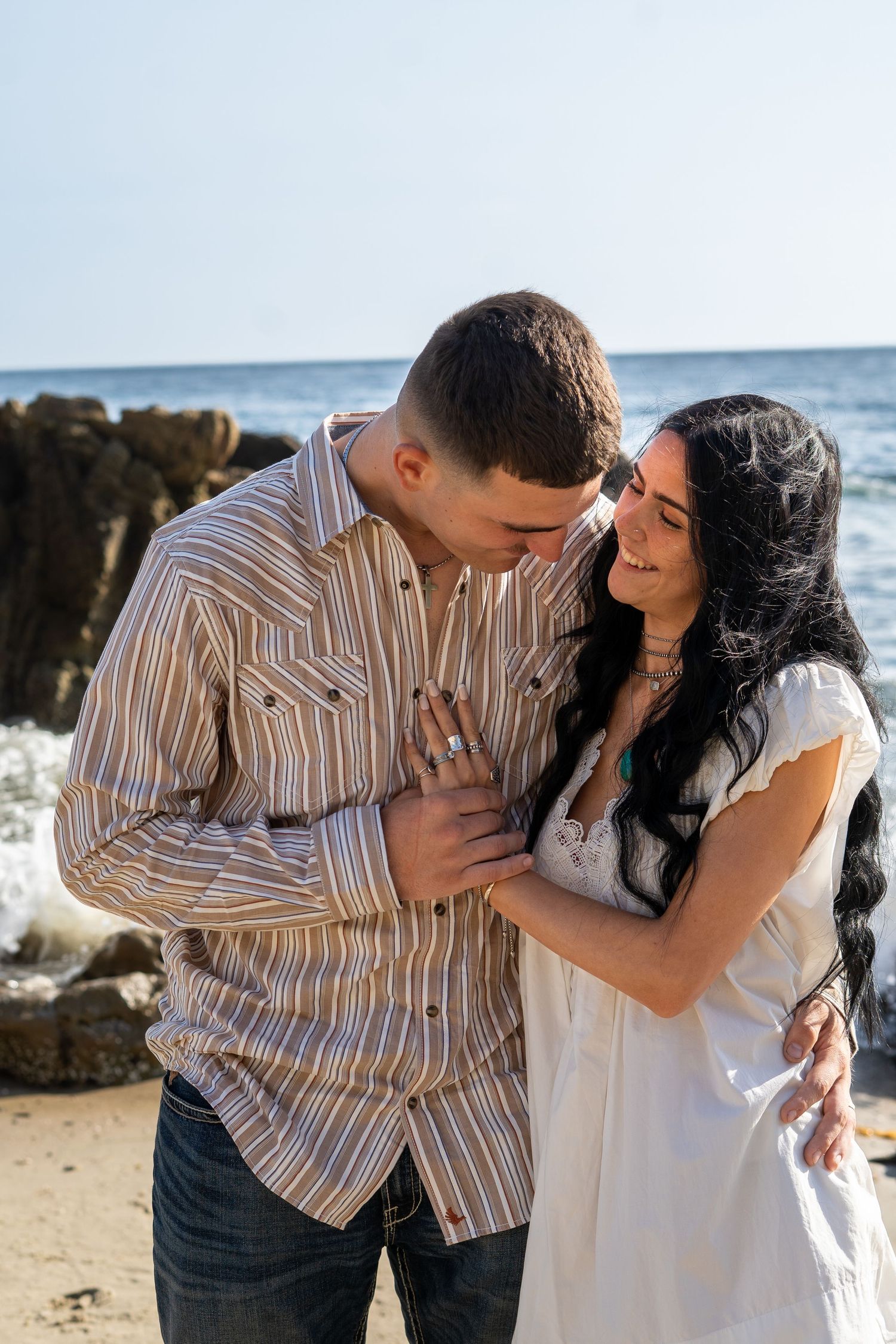 Couple embracing romantically on a rocky beach, woman in white dress and man in striped shirt, ocean waves in background.