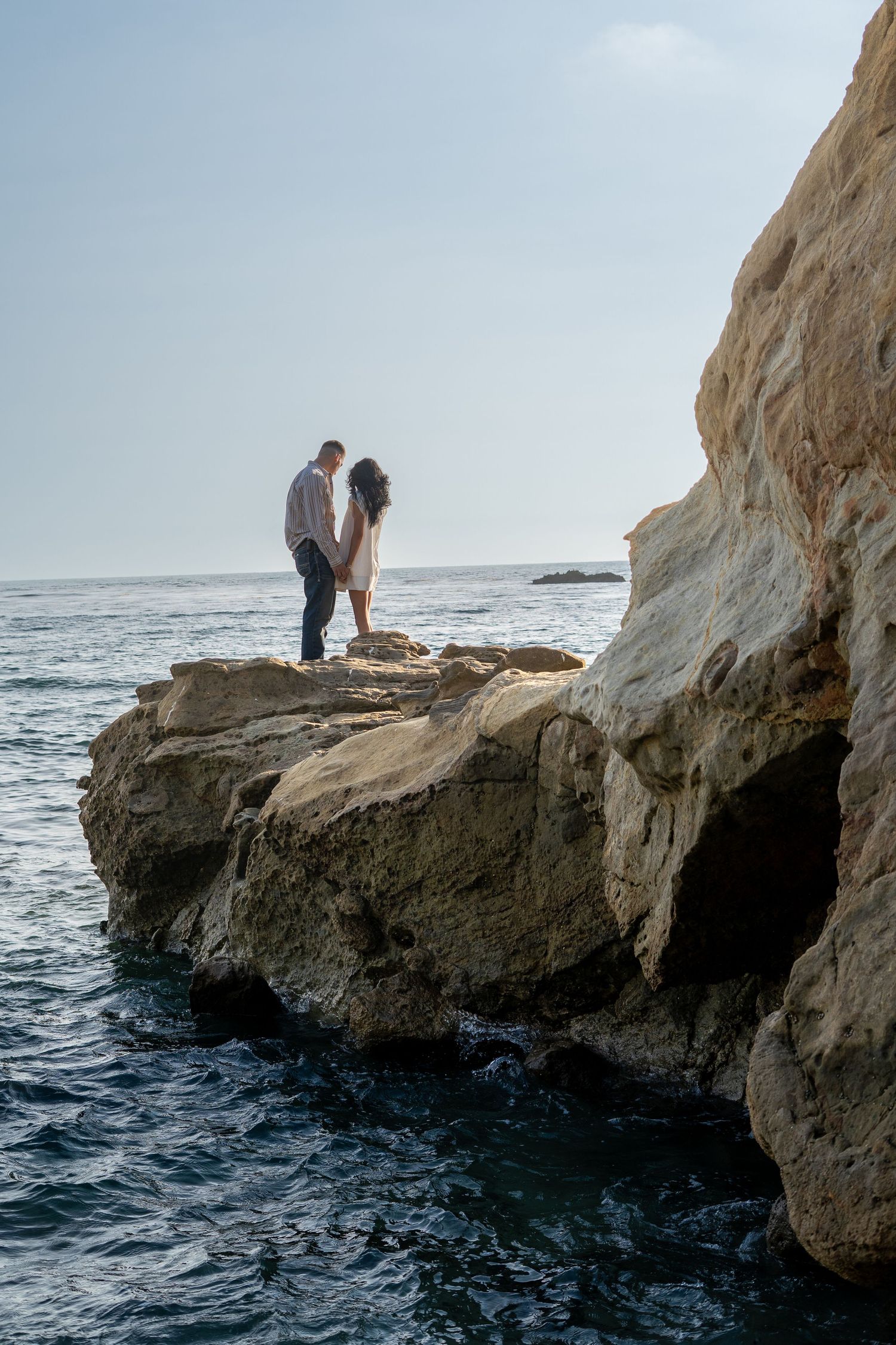 Romantic couple standing on rocky coastal cliffs above blue ocean waters during a sunny day.