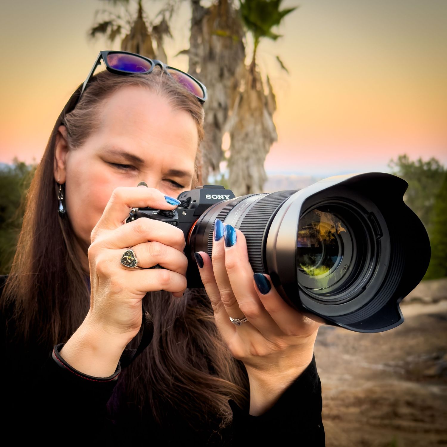 Woman photographer with blue nails photographing with Sony camera and large lens at sunset near palm trees.