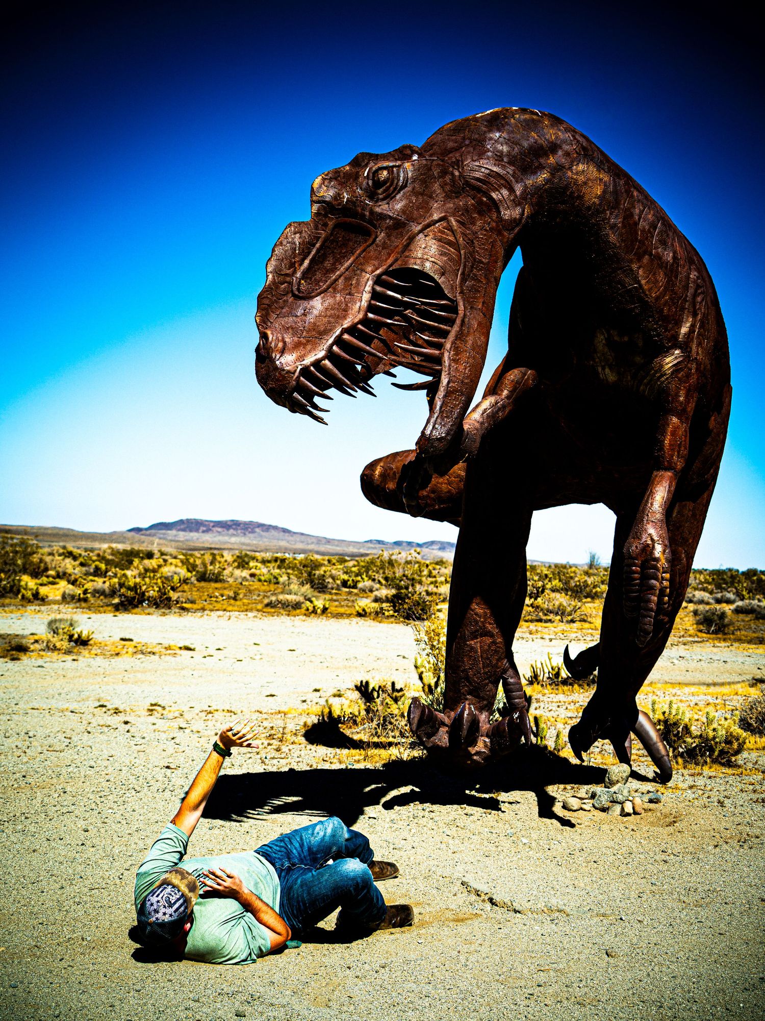 Person posing under a large T-Rex dinosaur statue in a desert landscape with clear blue sky.