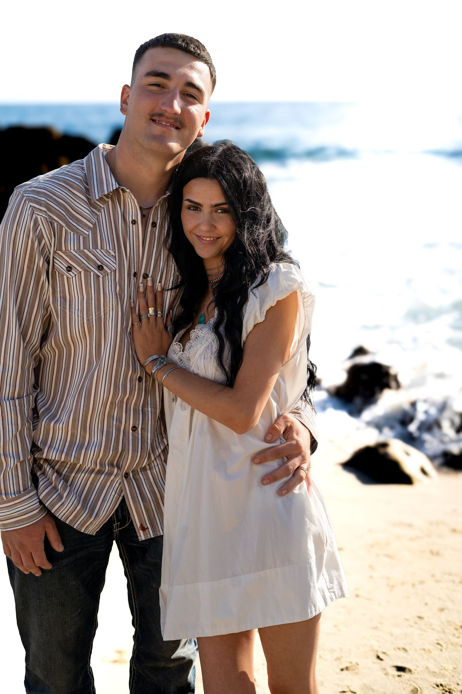 Young couple embracing on a sandy beach with ocean waves and rocks behind them.
