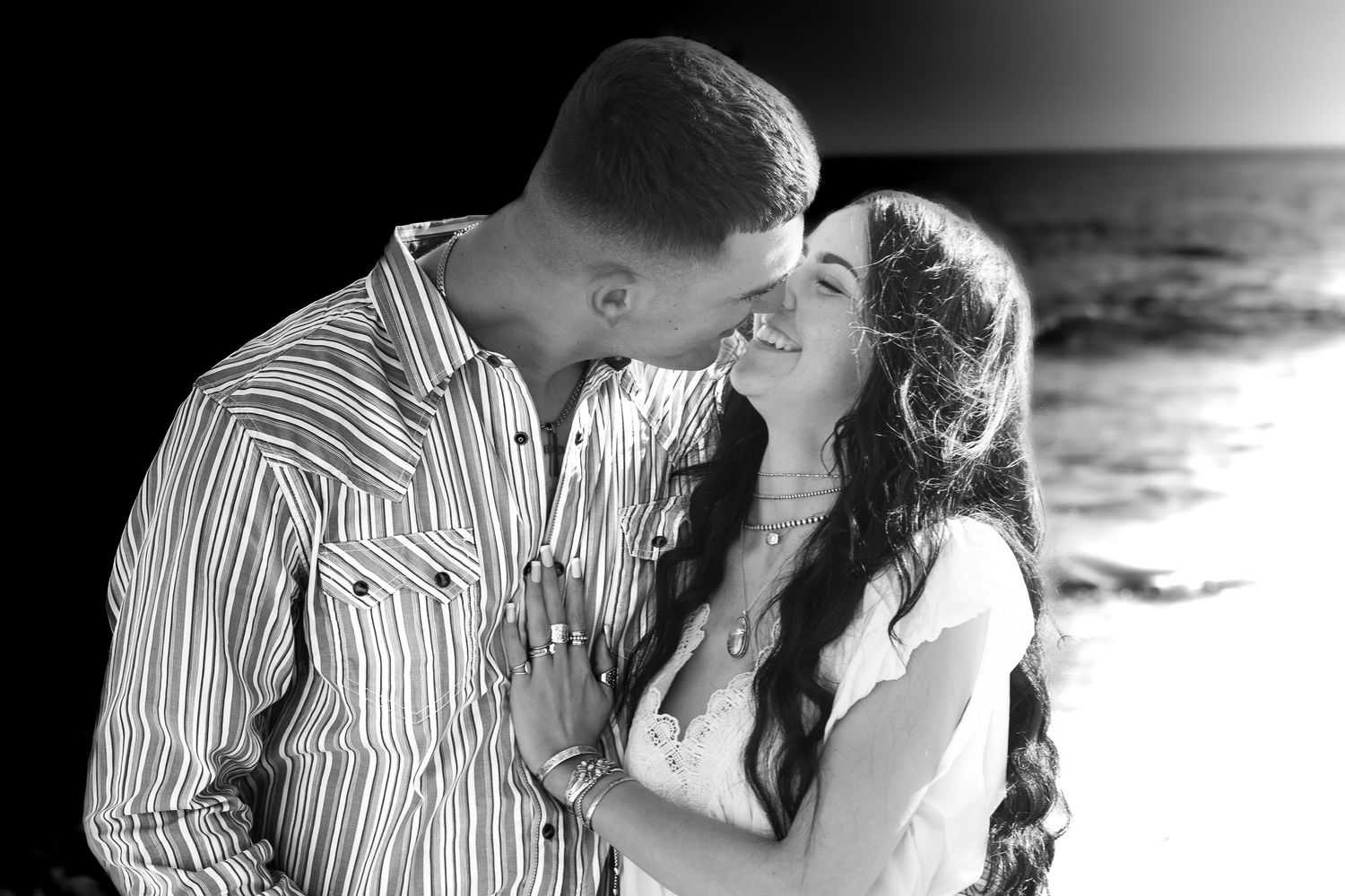 Black and white photo of a romantic couple smiling at each other on a beach with ocean waves in the background.
